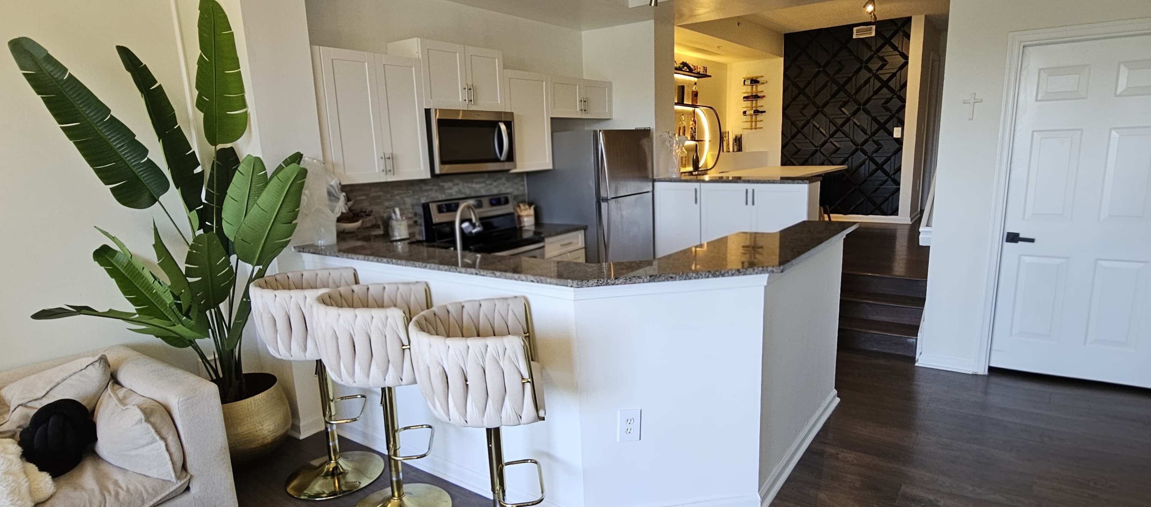 The image shows a modern kitchen with white cabinetry, stainless steel appliances, and a bar area with gold-accented stools, adjacent to a staircase and a seating area.