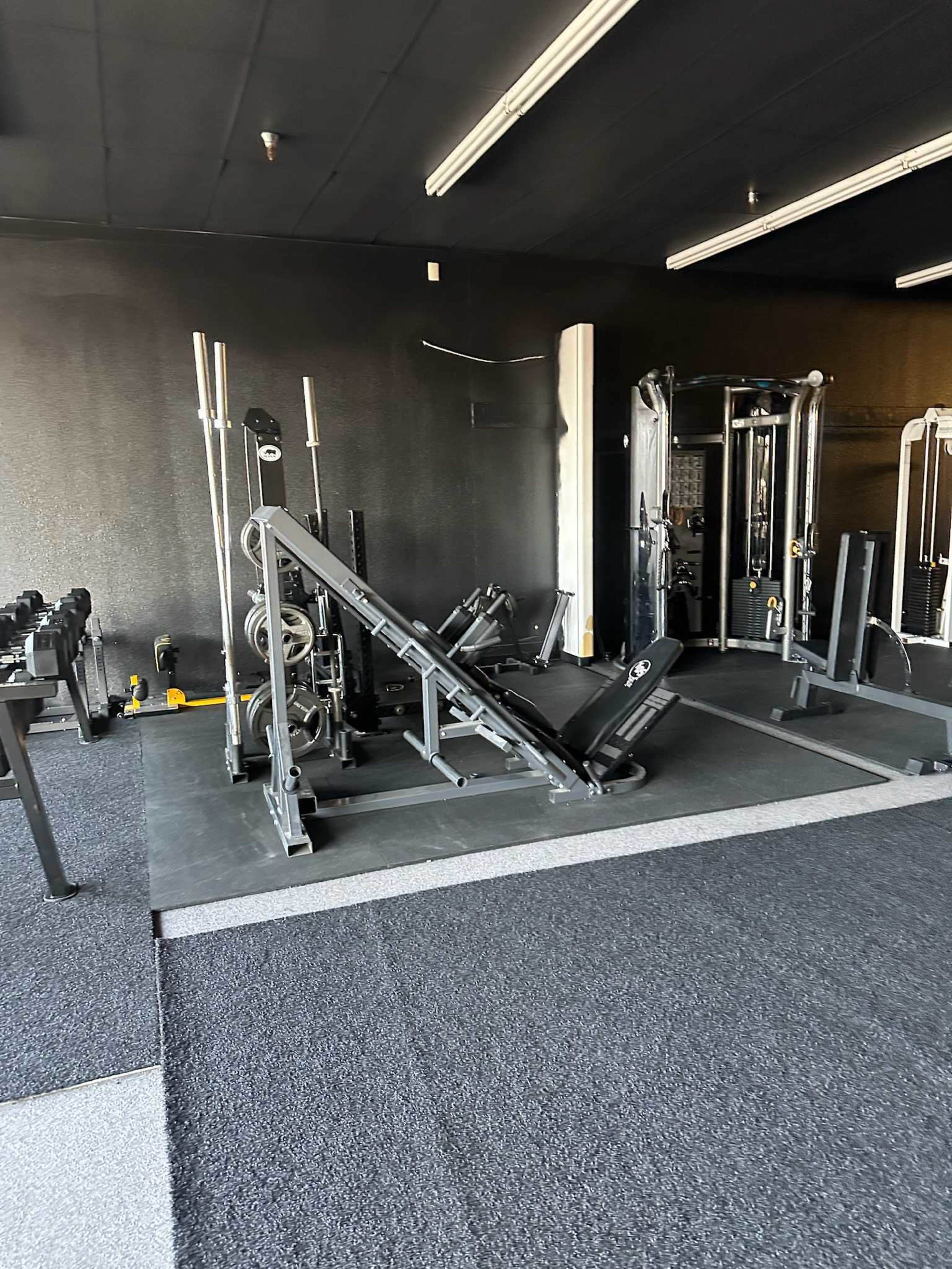 The image shows a gym interior featuring various weightlifting equipment, including a bench press and machines, on a black rubber flooring surface.