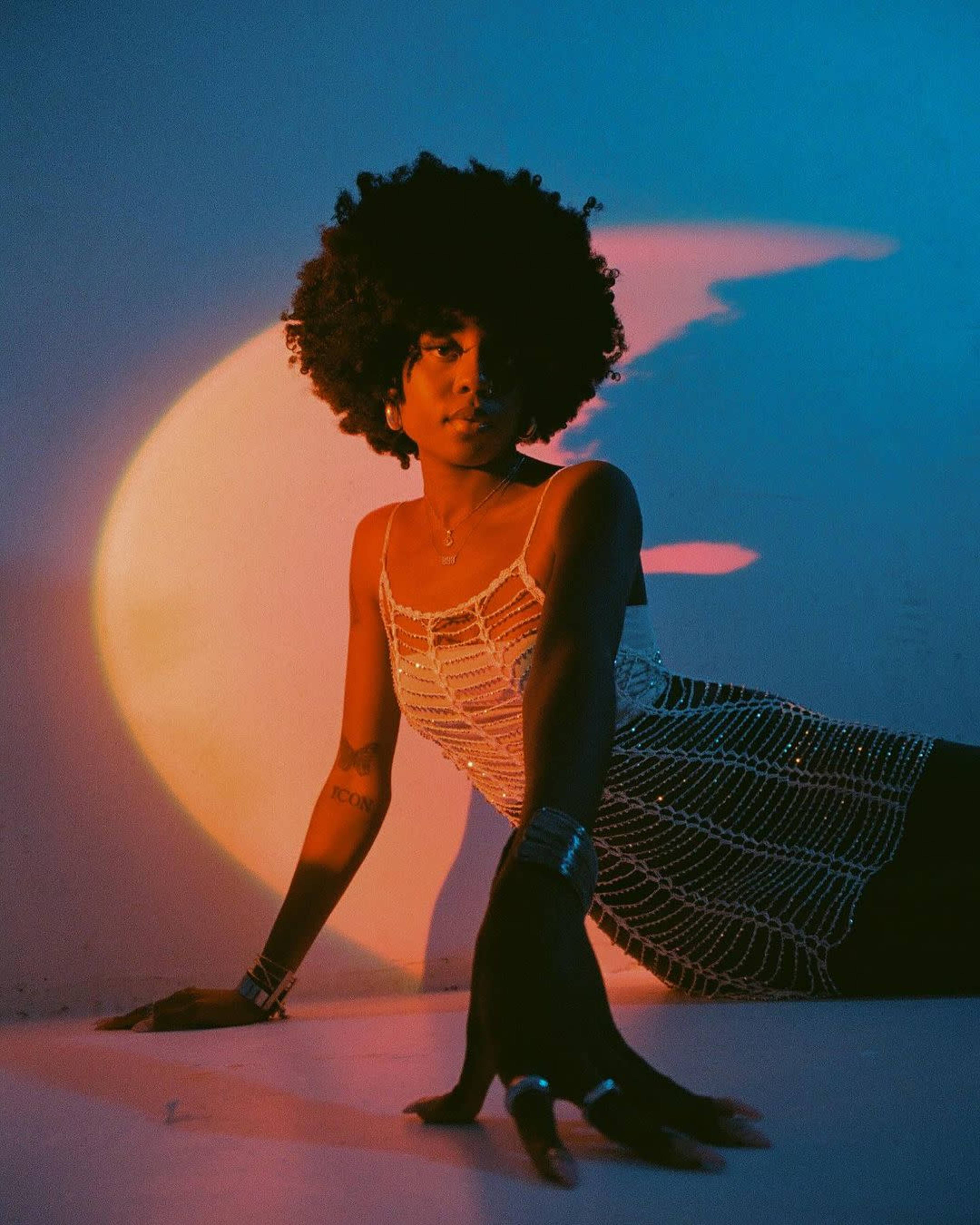 A woman with an afro hairstyle poses against a backdrop illuminated by colorful lighting.