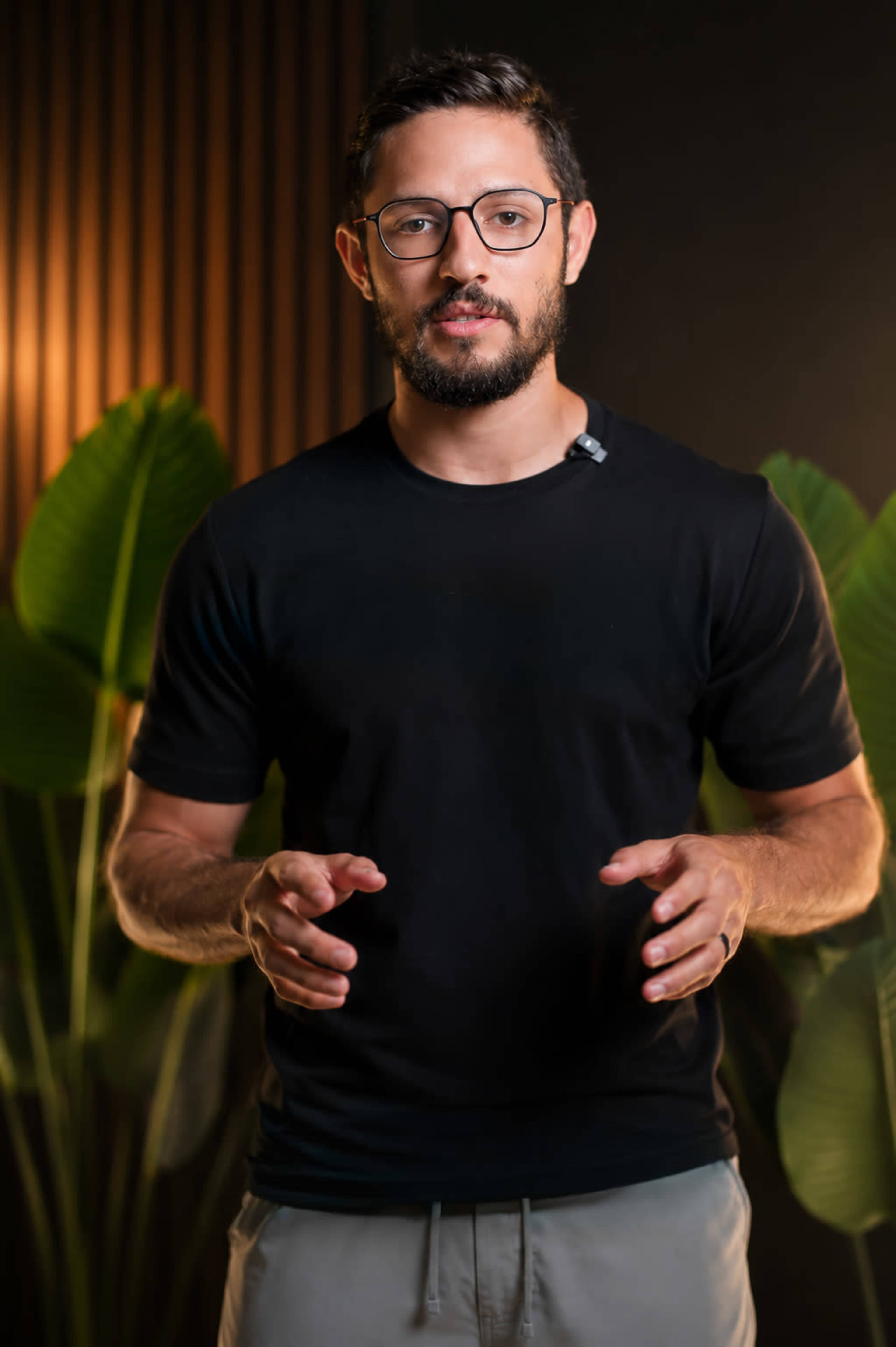 A man wearing glasses and a black t-shirt stands in front of a backdrop featuring large green plants.