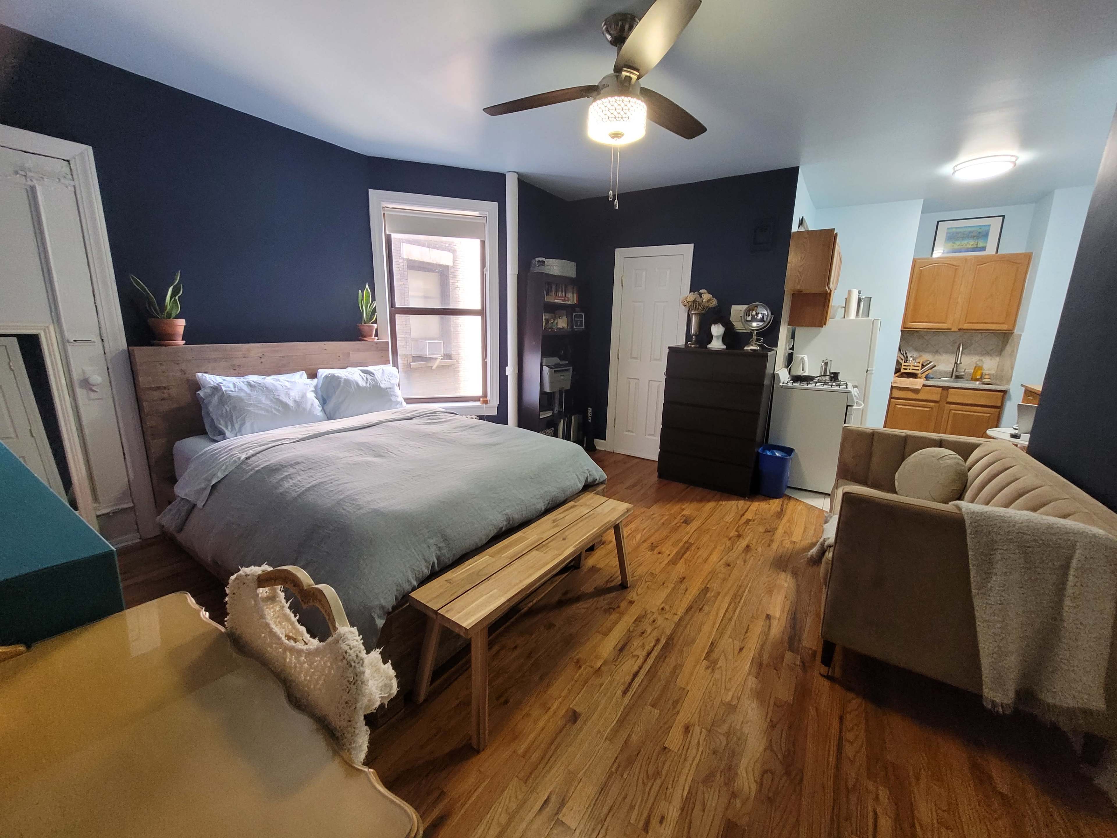 A well-furnished studio apartment featuring a bed, a seating area, a kitchenette, and hardwood floors.