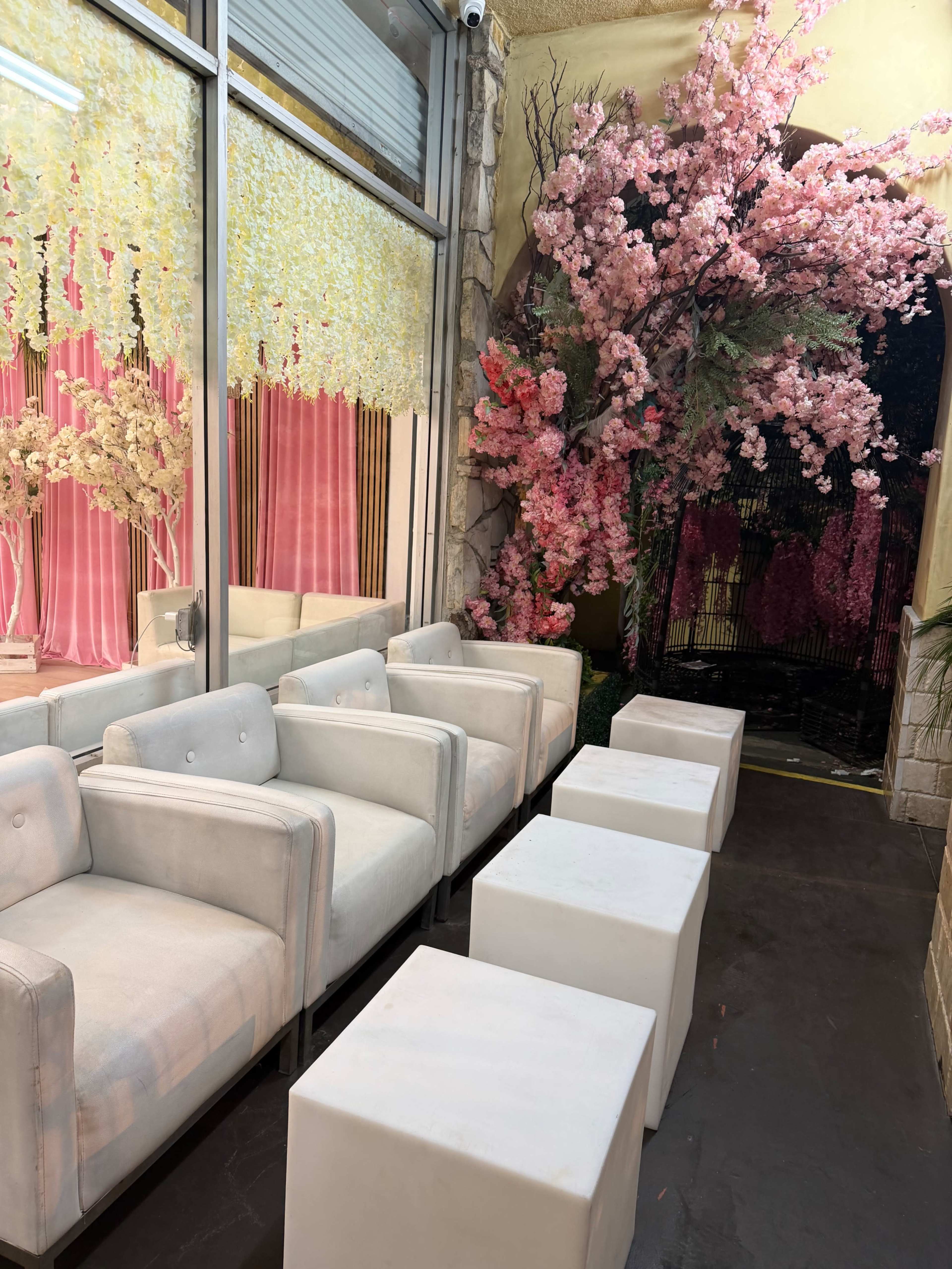 The image shows a cozy indoor space with white chairs and square tables, adorned with pink cherry blossom decorations and backlighting.