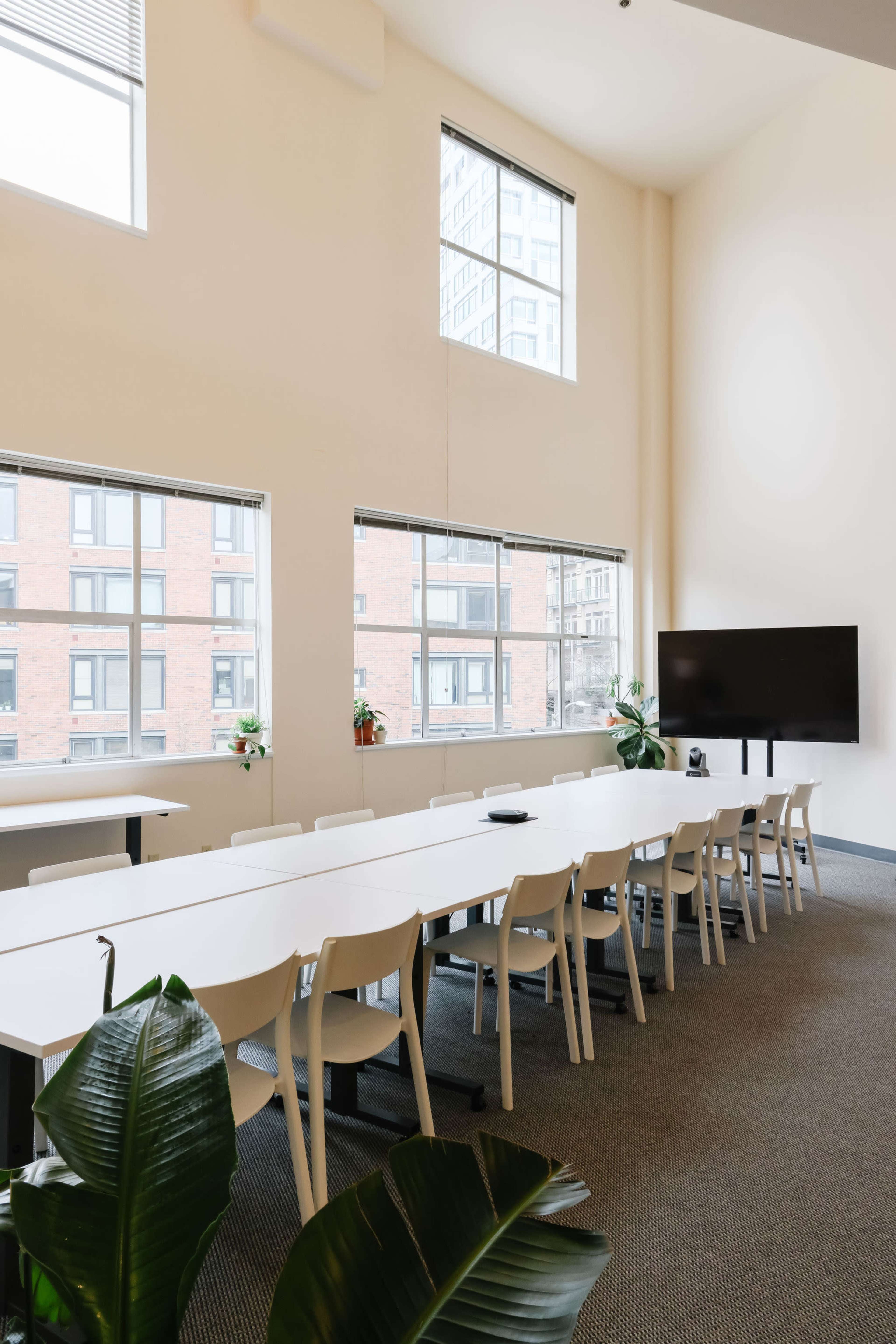 A bright conference room features a long white table surrounded by chairs, large windows, and a wall-mounted screen.