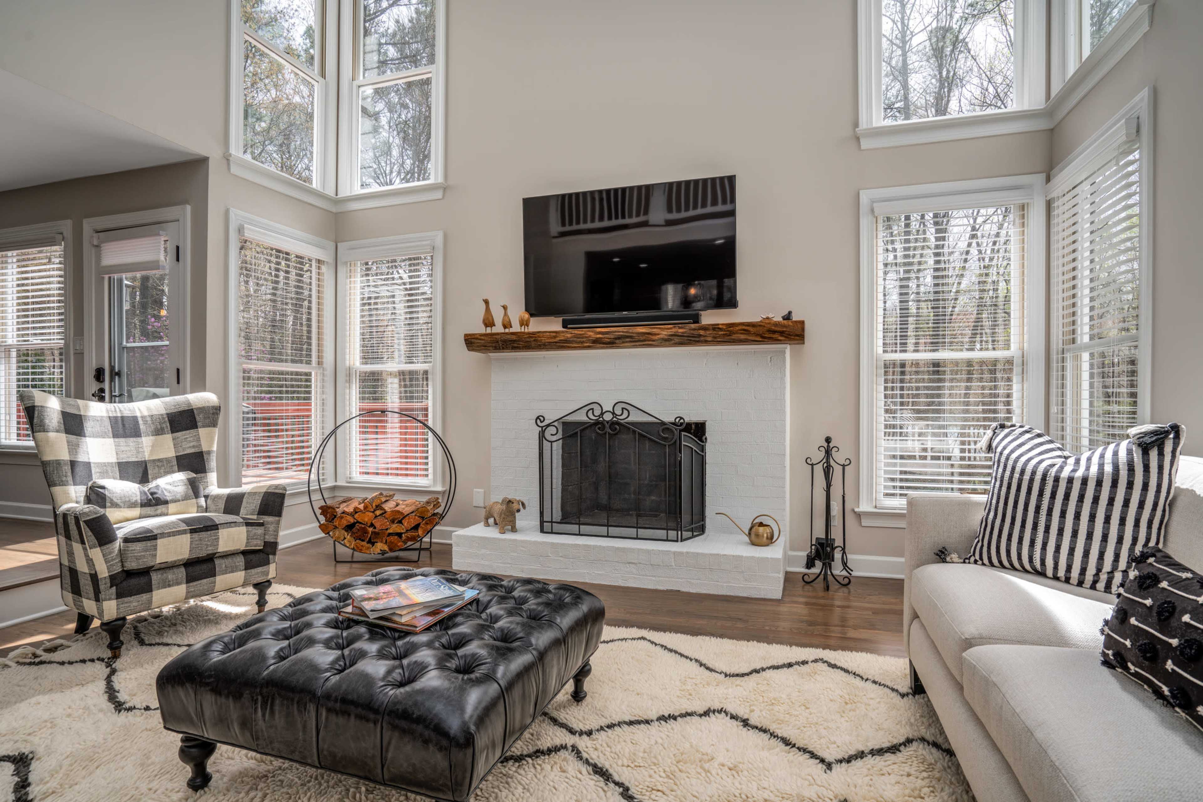 The living room features large windows, a white brick fireplace, a black tufted ottoman, and comfortable seating arranged around a central coffee table.