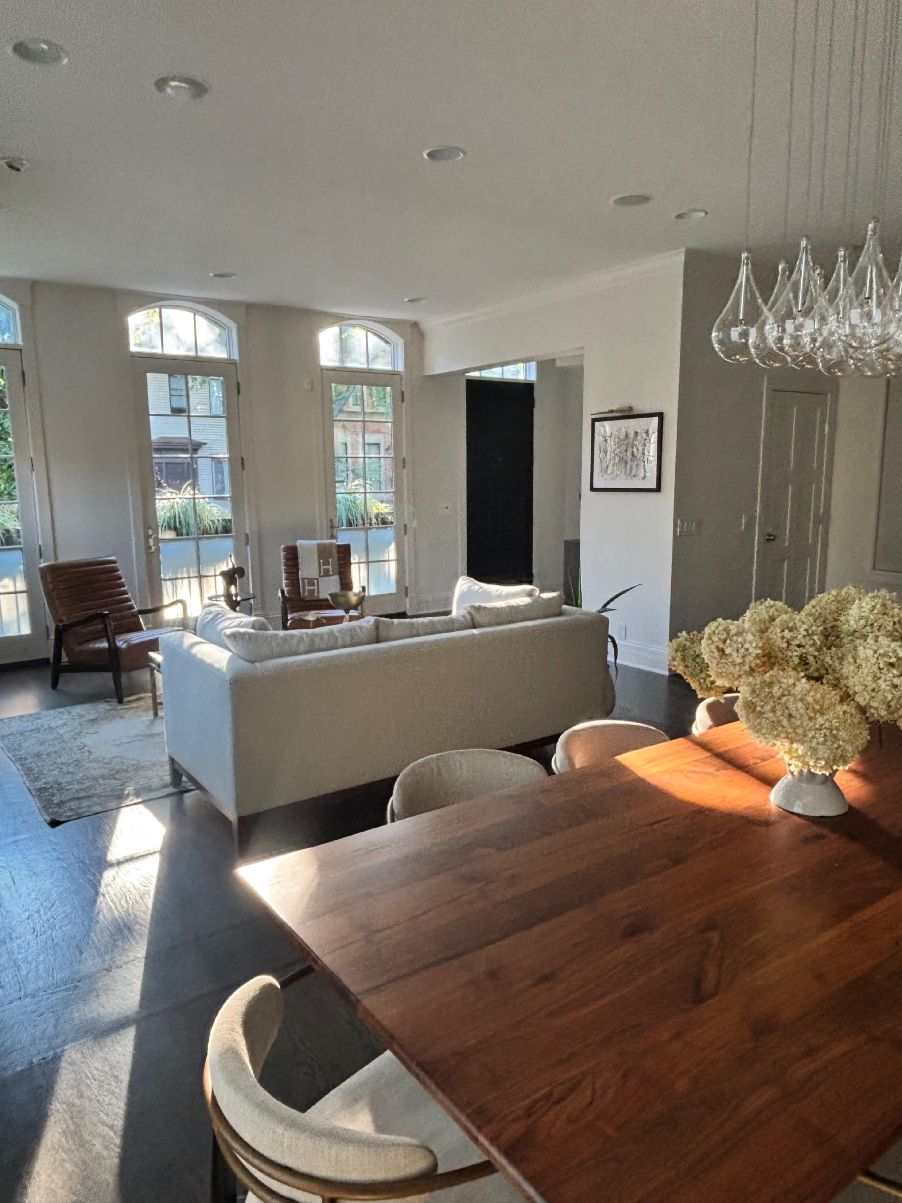The image shows a modern living space with a light-colored sofa, a wooden dining table, and large windows allowing natural light to fill the room.