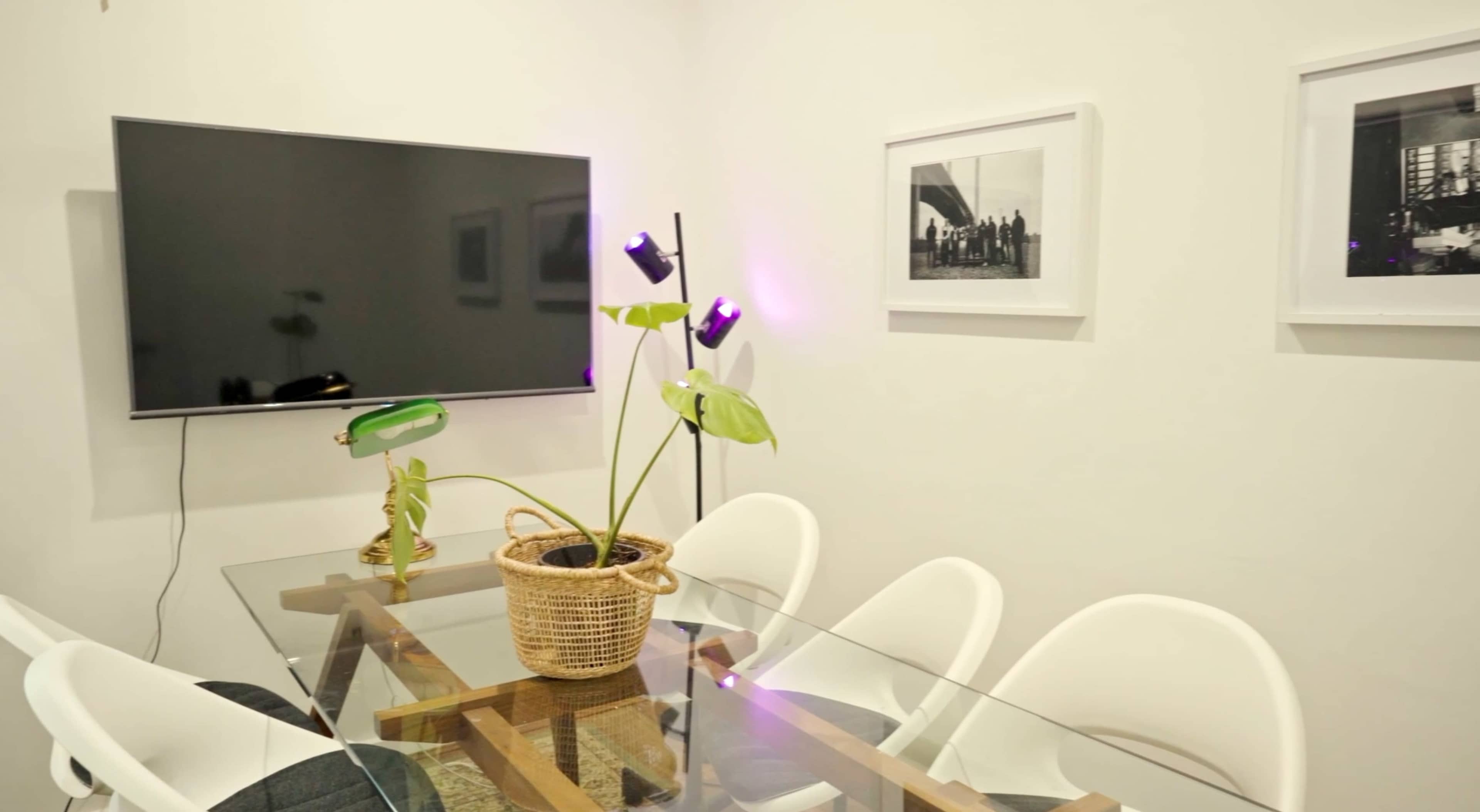 A glass dining table with white chairs is positioned under a wall-mounted television, alongside a potted plant and framed photographs on the walls.