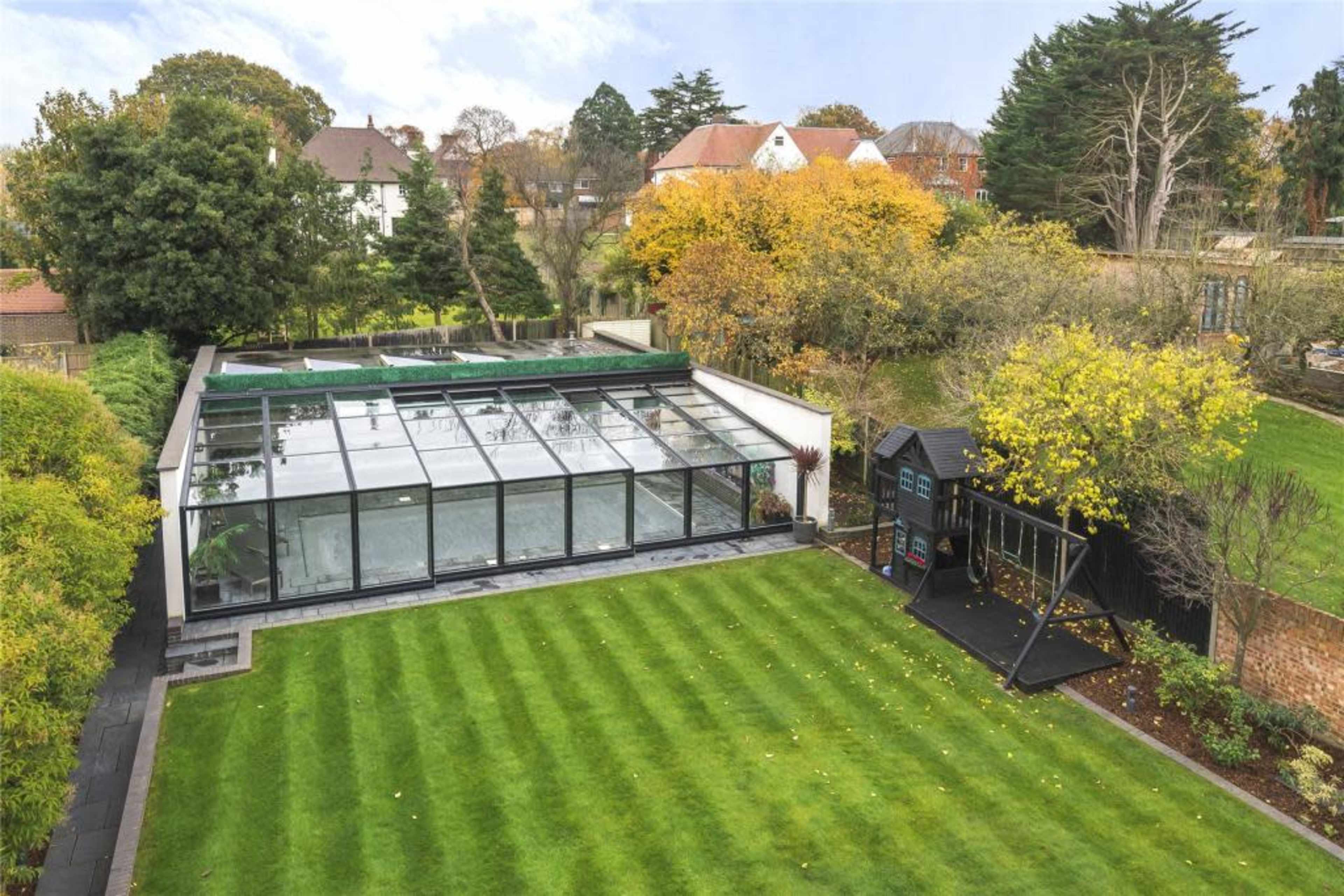The image shows a modern glass extension attached to a house, set in a well-maintained lawn surrounded by trees and autumn foliage.