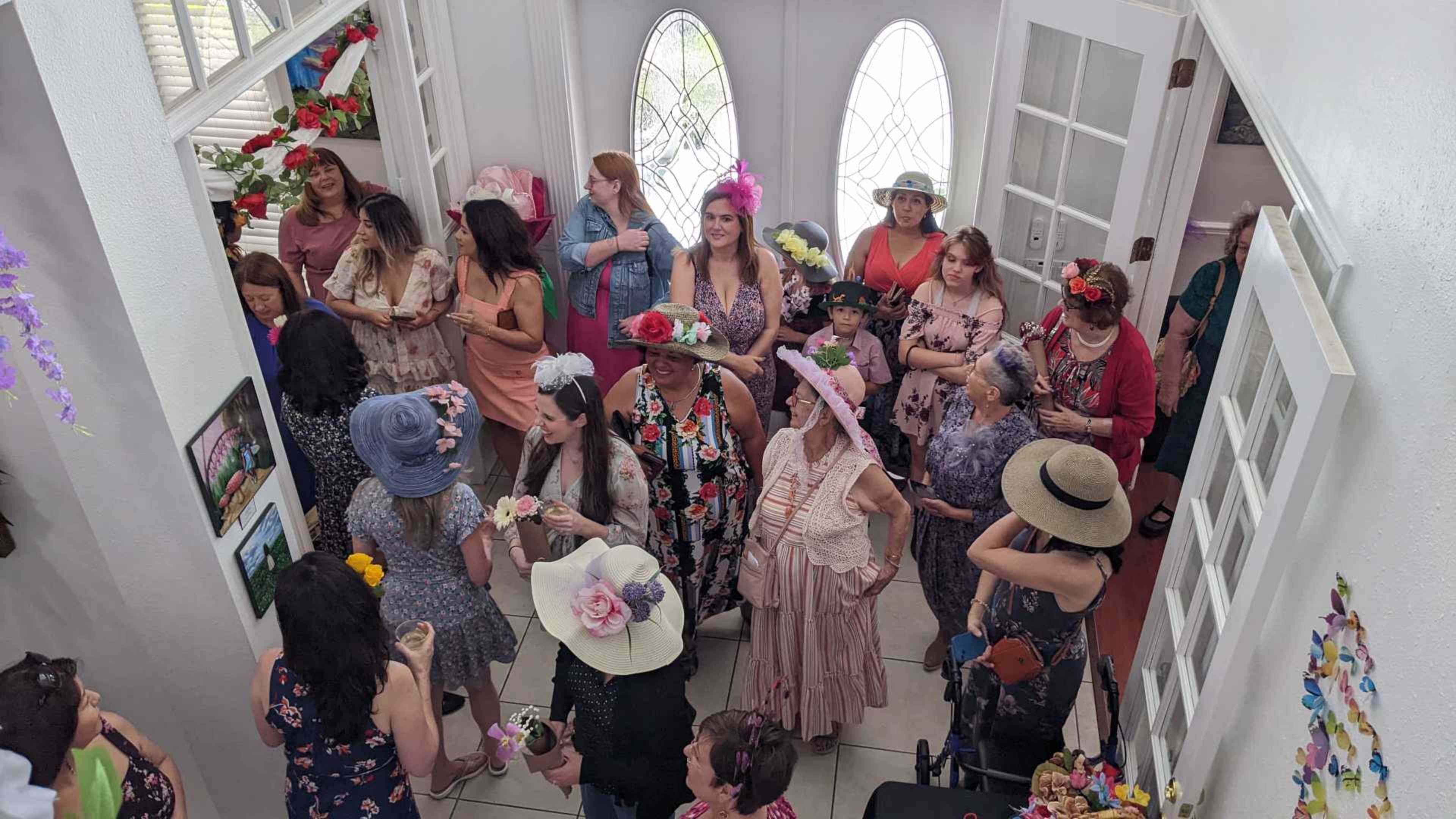A group of women, many wearing decorated hats, gather in a brightly lit foyer adorned with flowers and photographs.