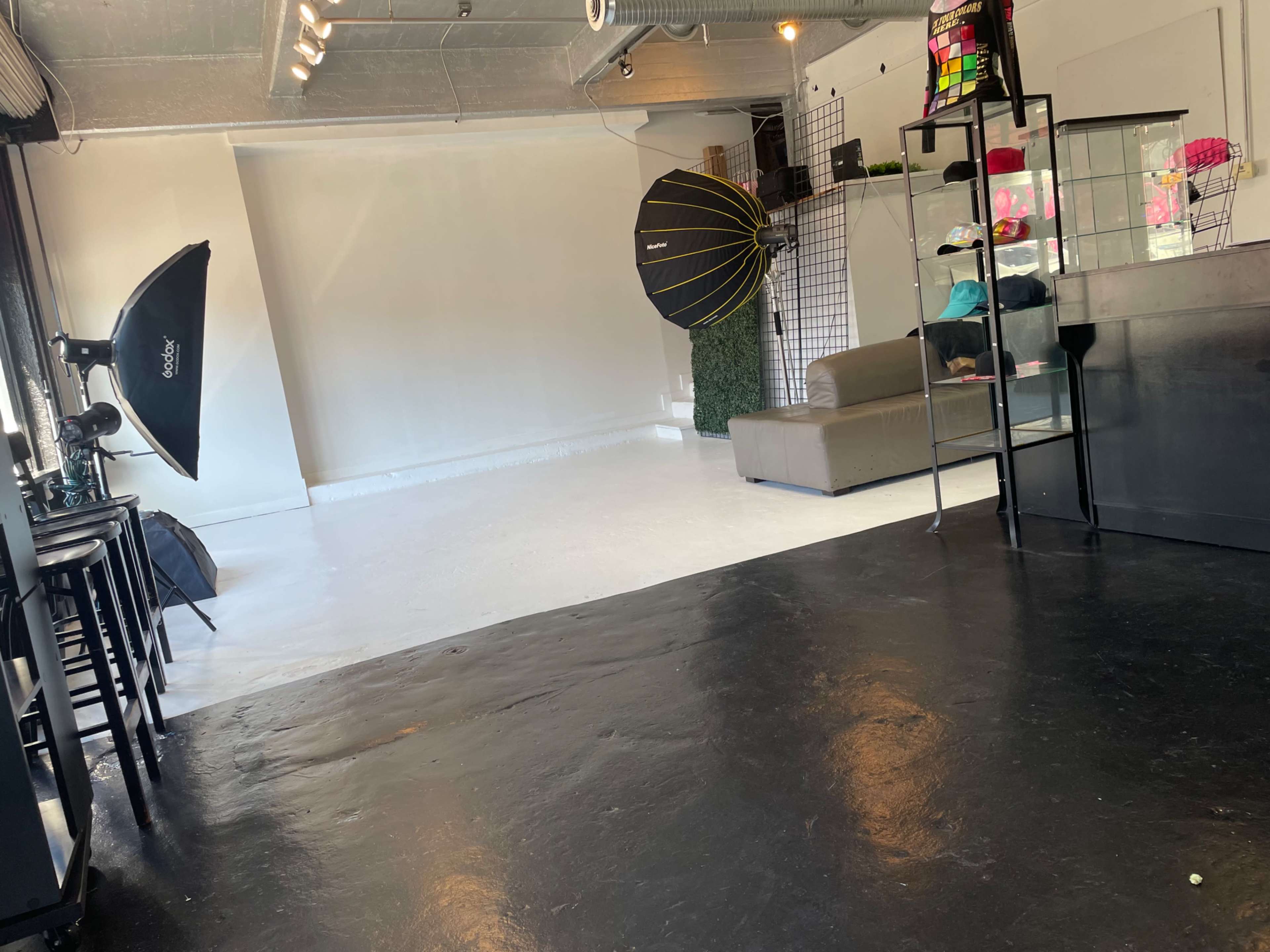 A spacious, minimalistic studio with black flooring, white walls, a gray sofa, and photography equipment including softboxes and a display case.