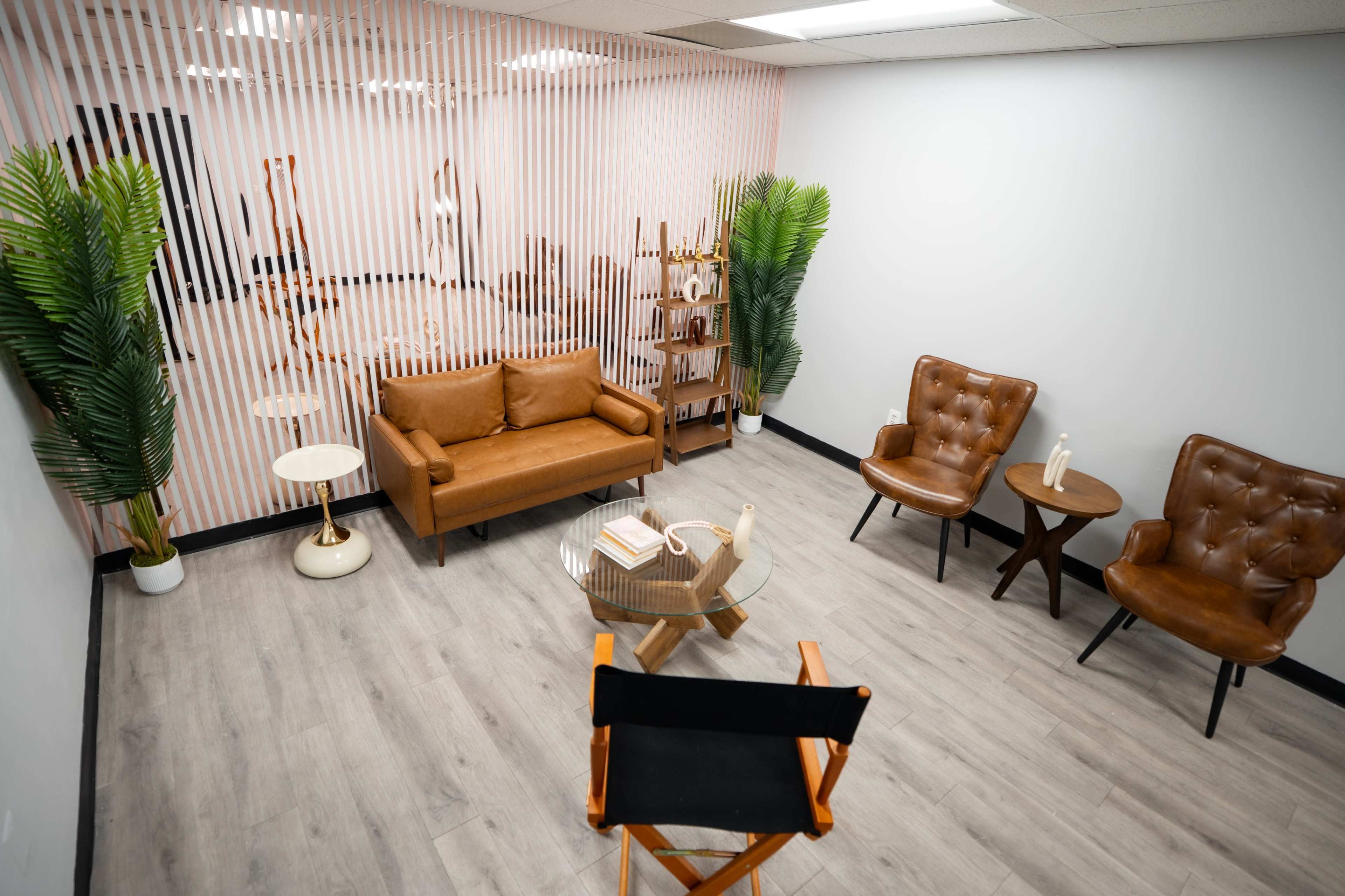 The image shows a modern waiting room with a brown sofa, two armchairs, a glass coffee table, and decorative plants.