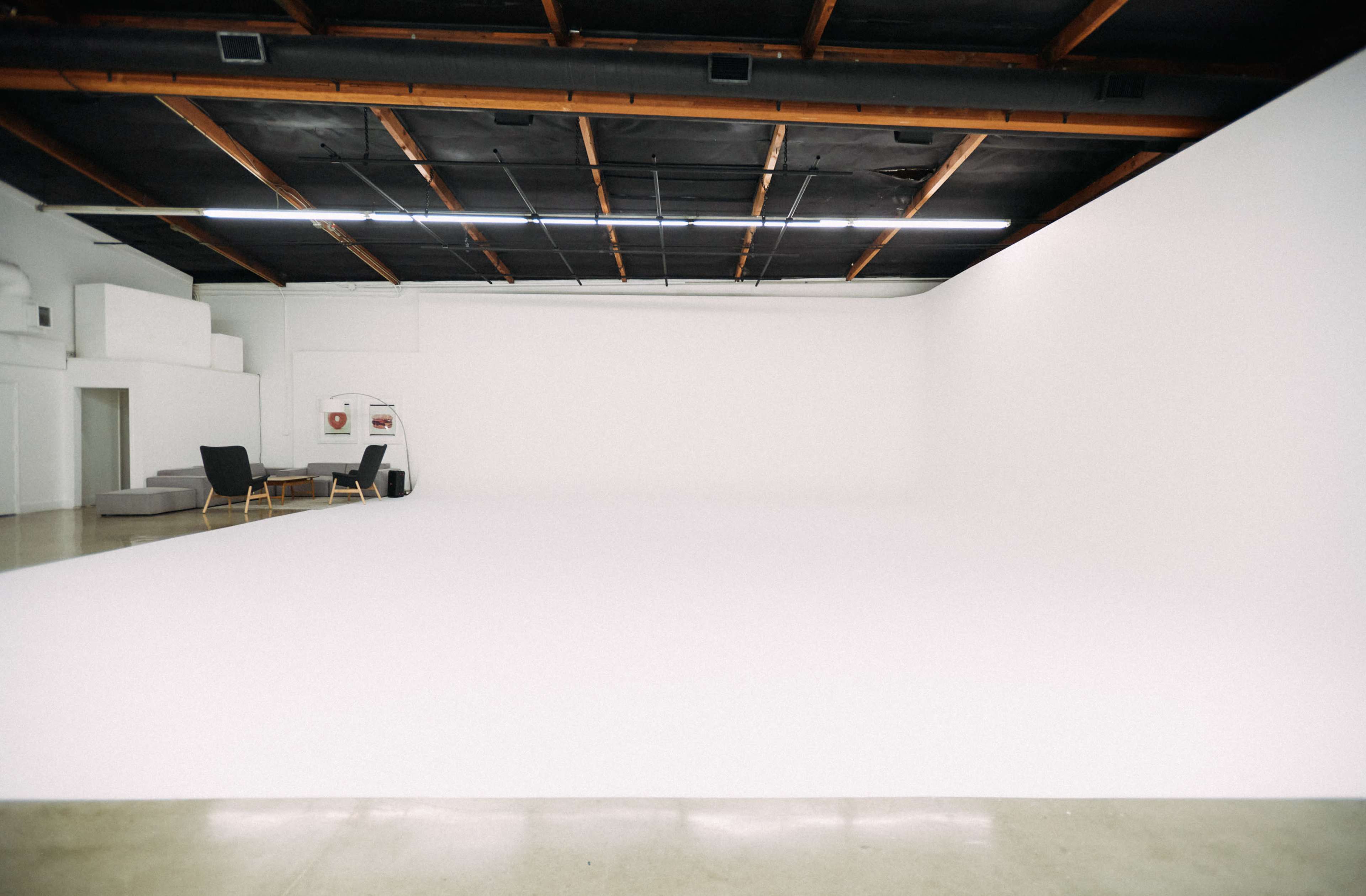 A spacious, empty studio with white walls, a smooth floor, and minimal furniture arranged in one corner.