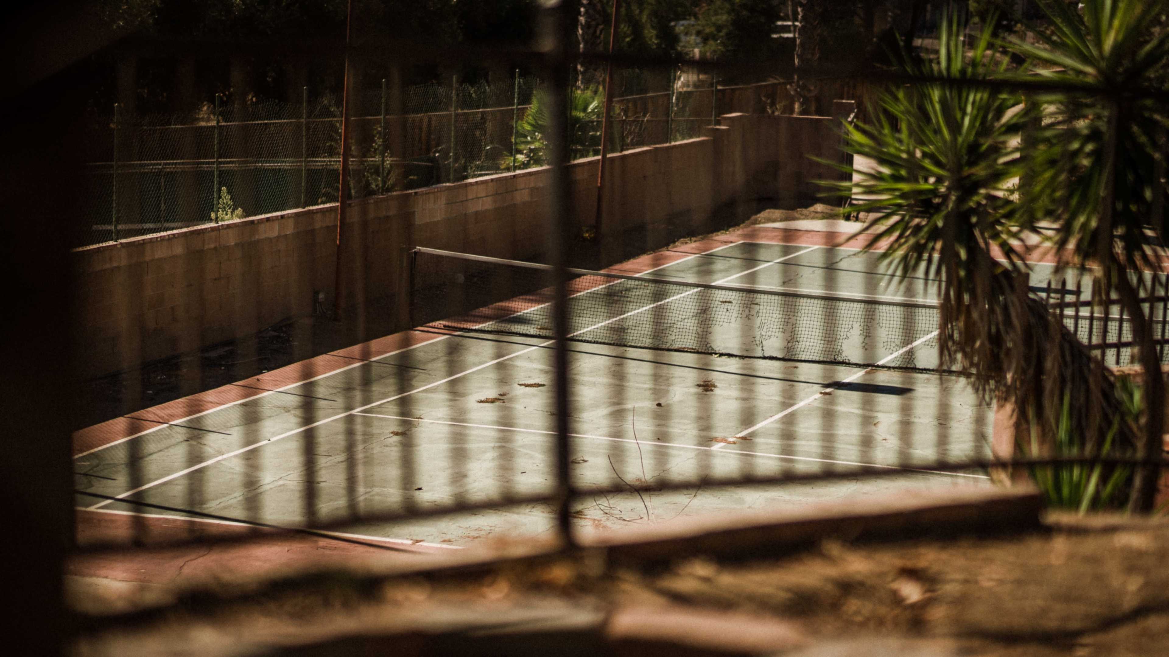 An abandoned tennis court with overgrown vegetation and debris on the surface is visible through a fence.