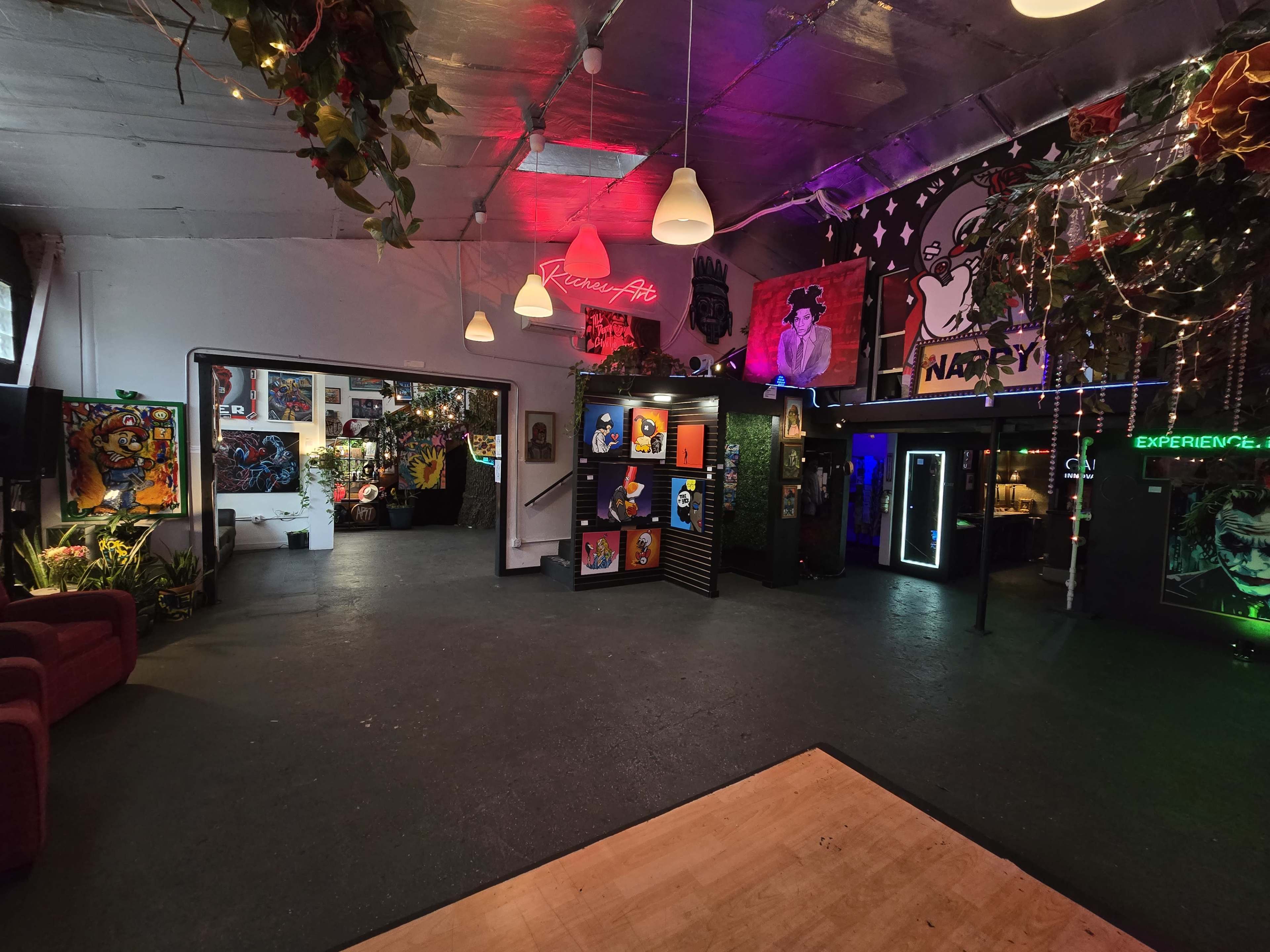 The image shows a vibrant indoor space decorated with colorful art, neon lights, and plants, featuring couches and various art installations along the walls.