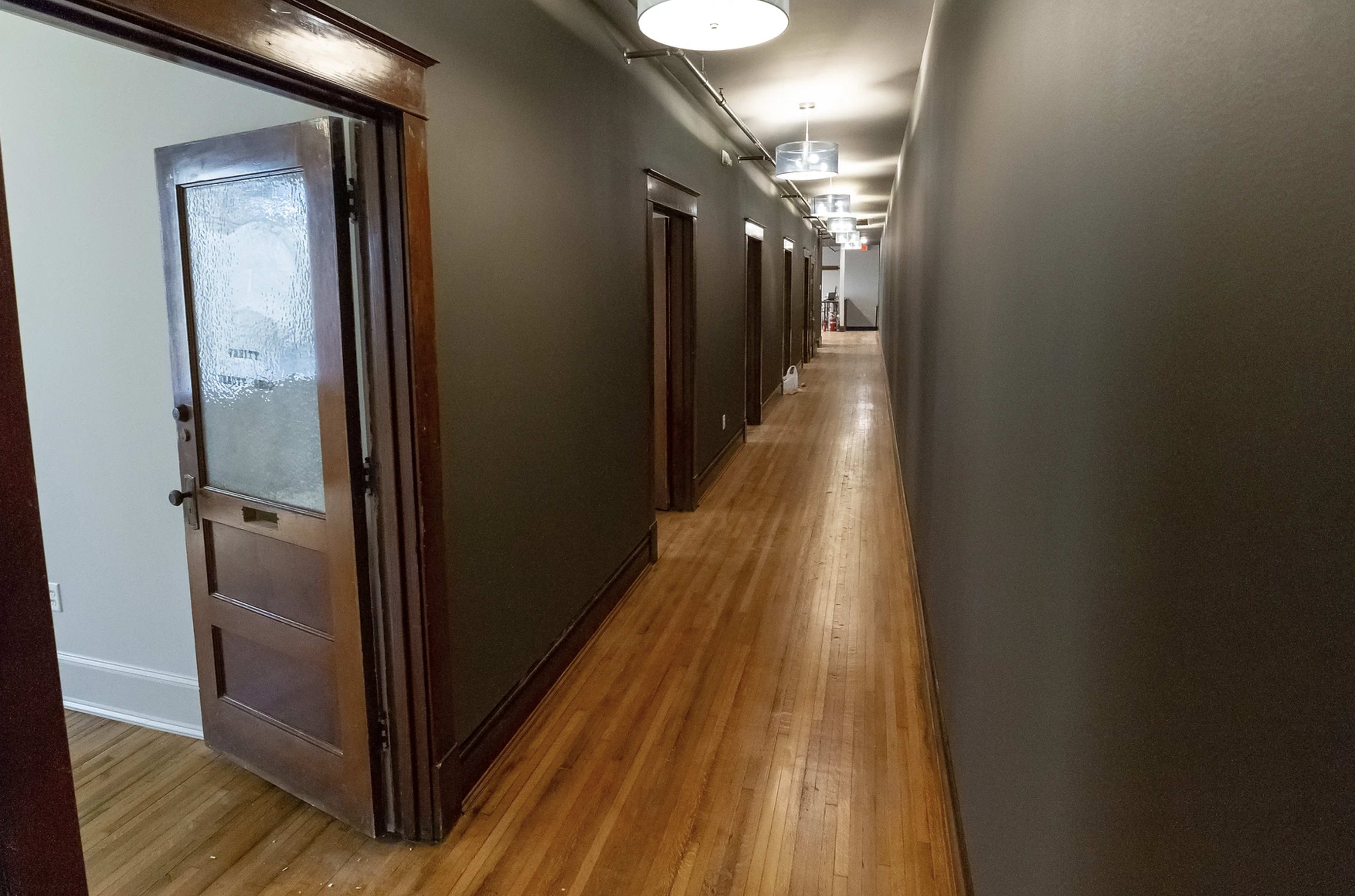 A long hallway with wooden flooring and several doors on either side, illuminated by overhead lights.