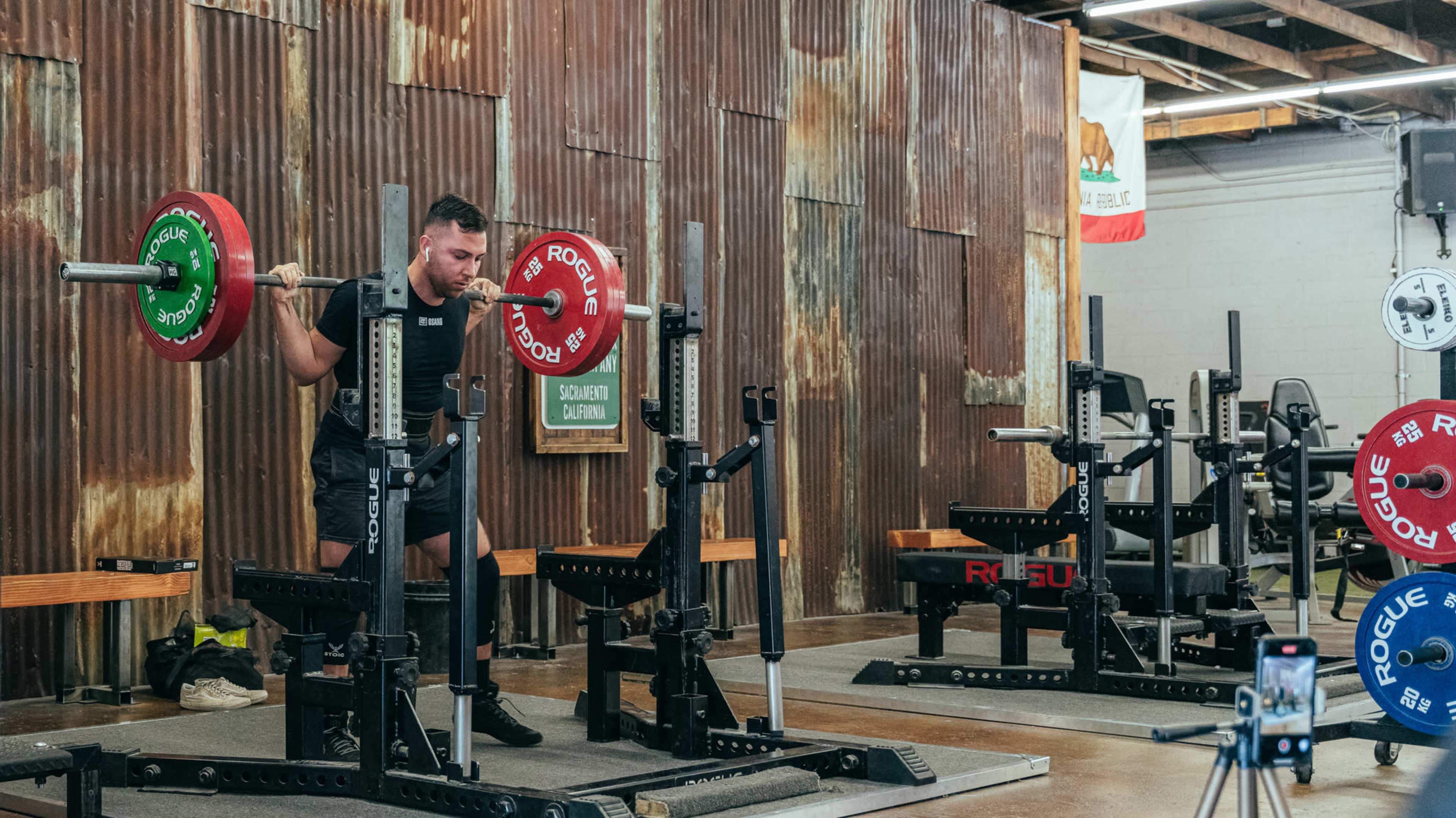 A person is performing a squat with a barbell loaded with weights in a gym setting with metal walls and equipment.