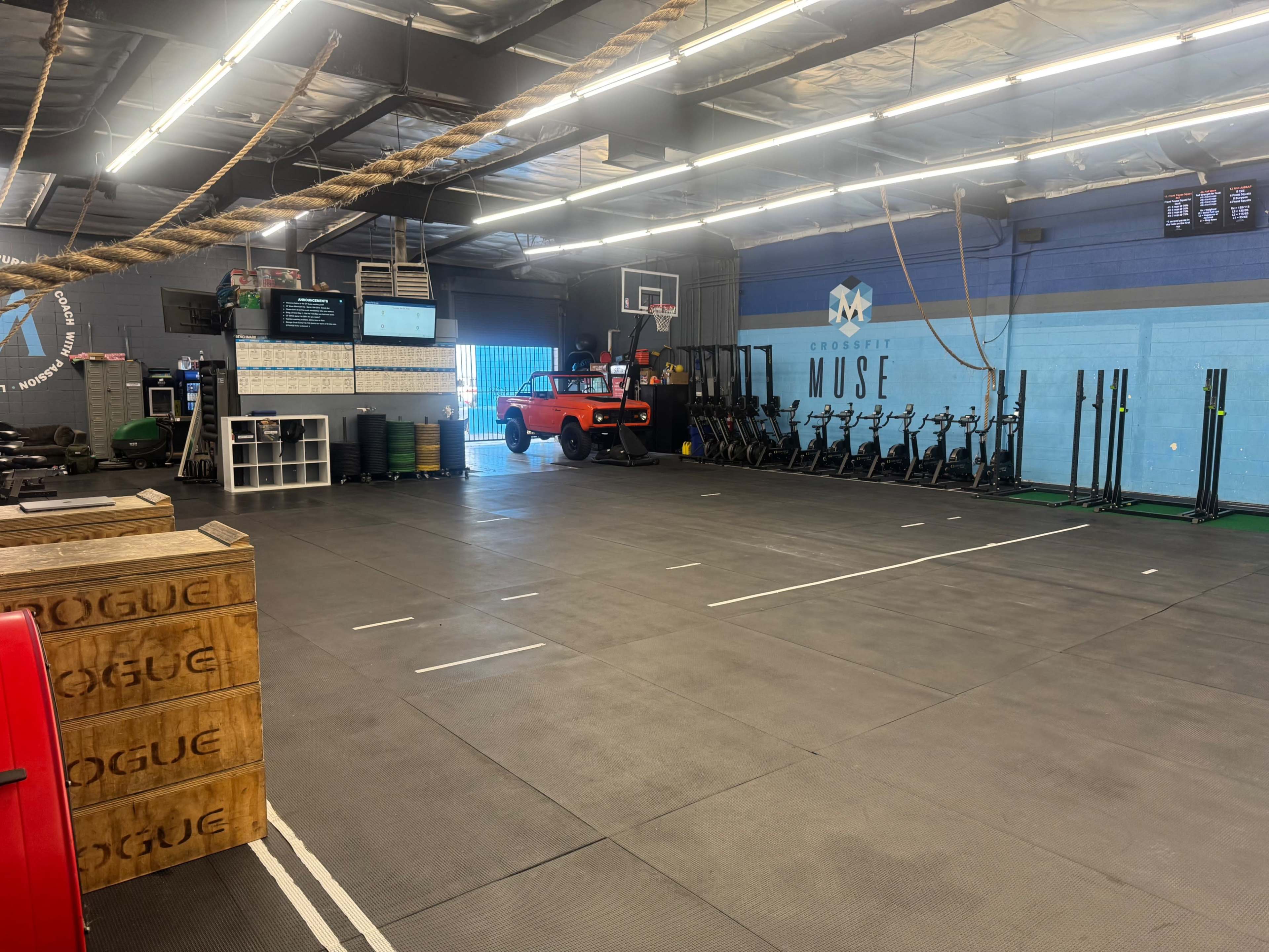 The image shows an empty gym space with exercise equipment, including rowing machines and weights, and a bright blue doorway leading outside.