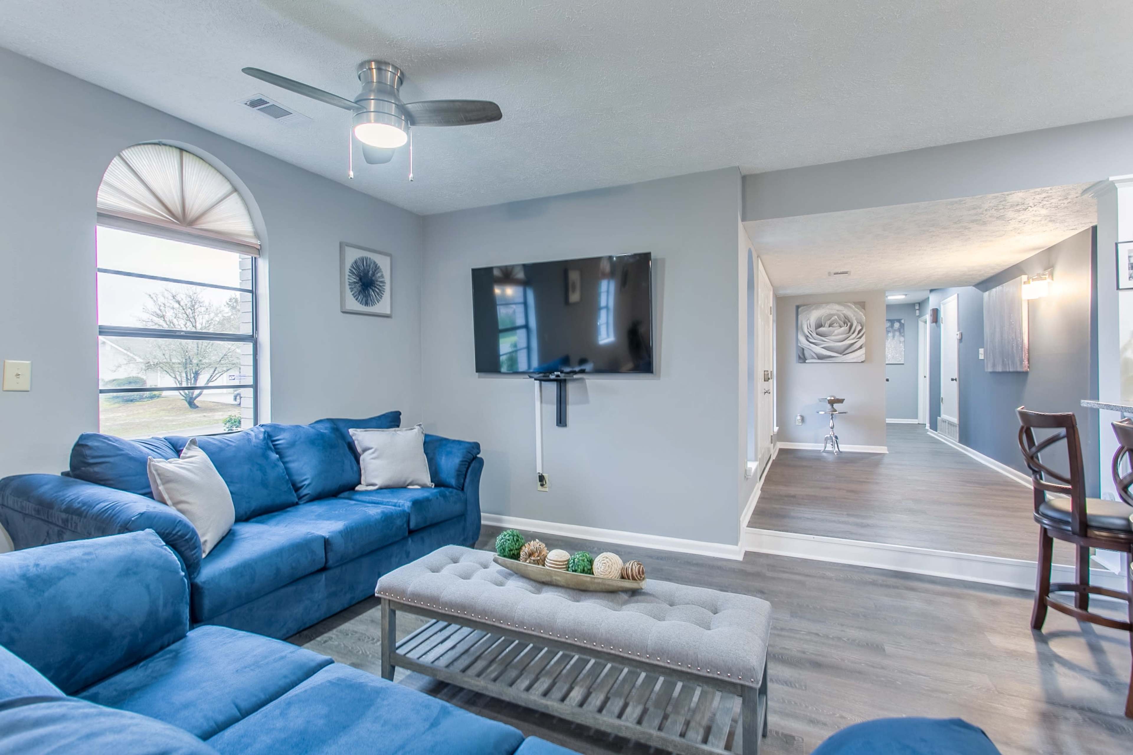 A living room with a blue couch, a wall-mounted television, and a view of an open hallway leading to another area of the house.