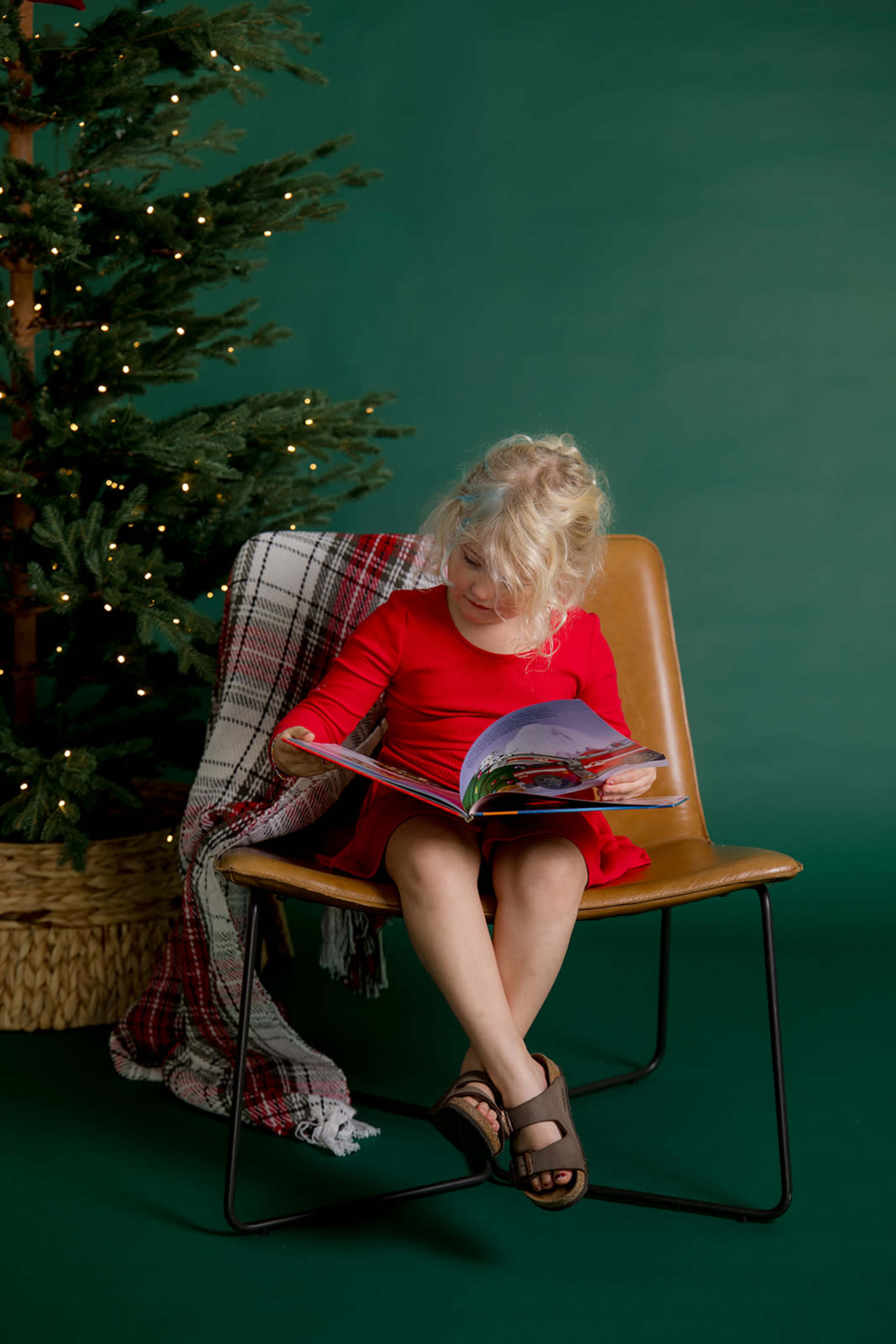 A young child sits on a chair, reading a colorful book in front of a decorated Christmas tree.