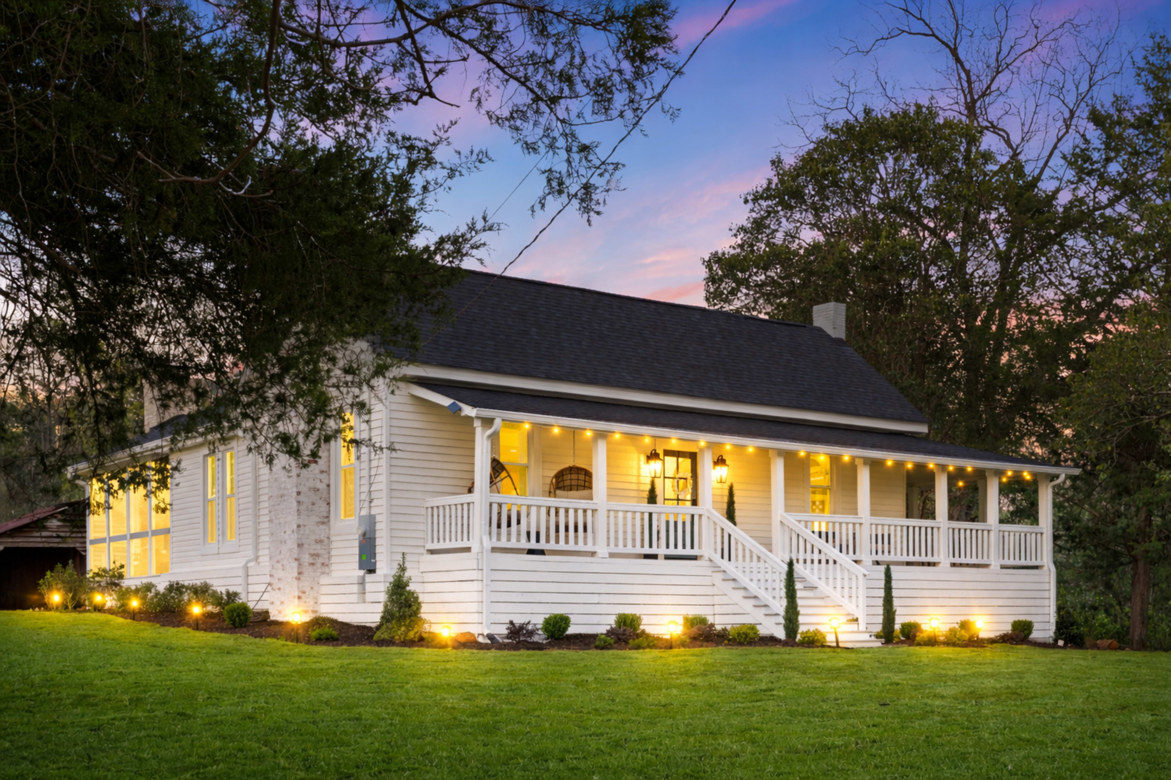 A white, one-story house with a front porch and landscaped yard is illuminated by outdoor lights at dusk.