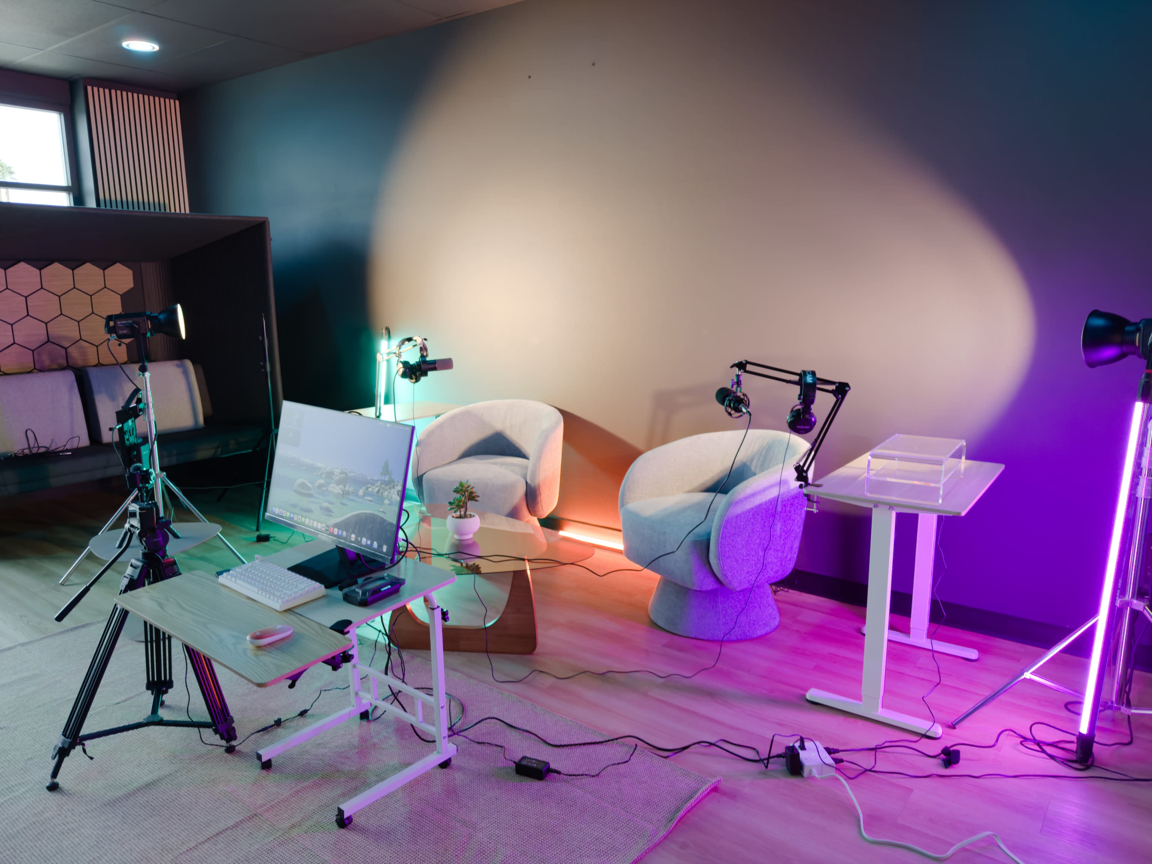 A modern filming setup features two light-colored chairs, a desk with a monitor, various cameras, and colorful lighting against a dark wall.
