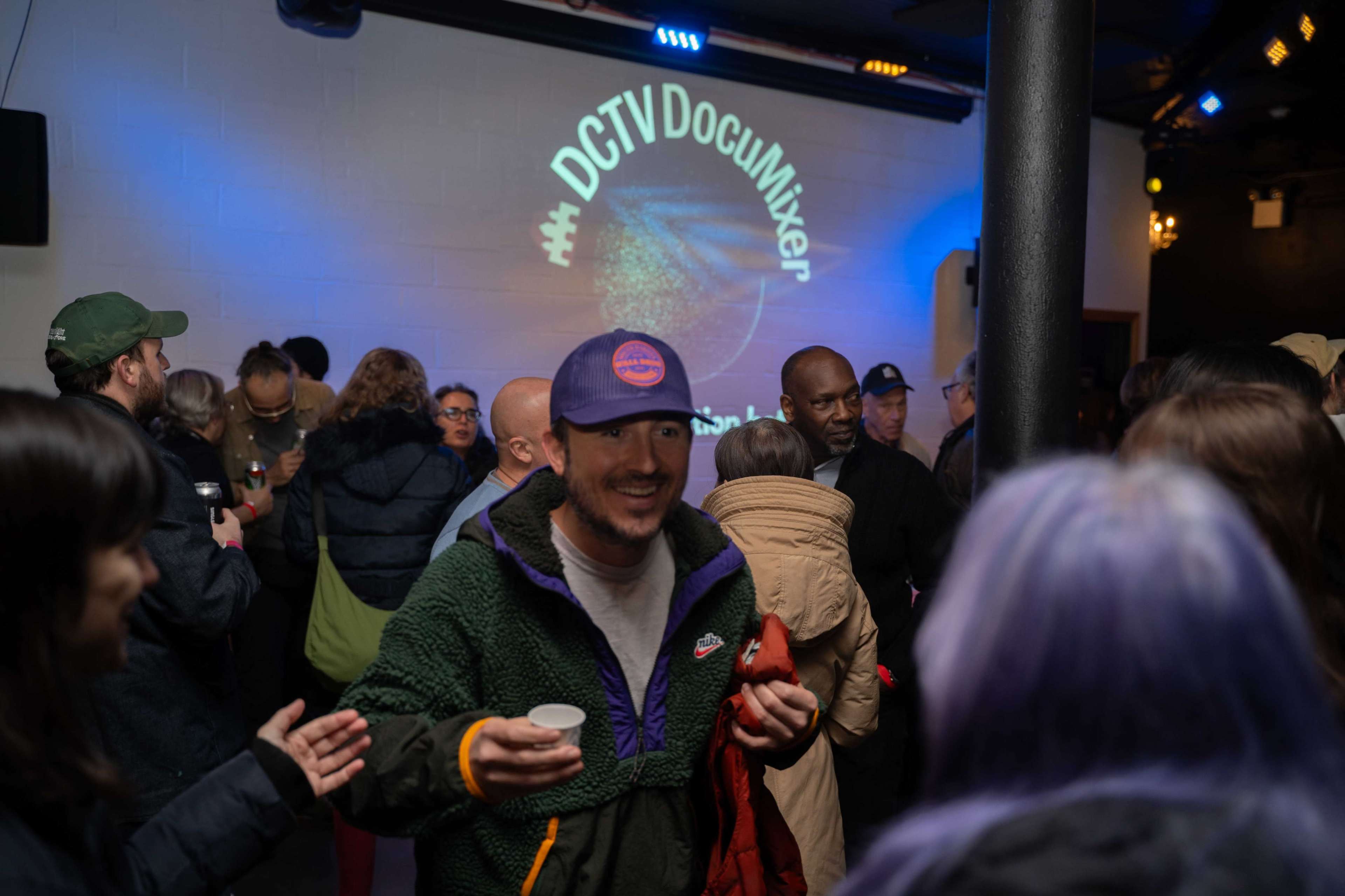 A crowd of people mingles in a dimly lit venue, with a projection displaying "#DCTVDocMixer" on the wall.