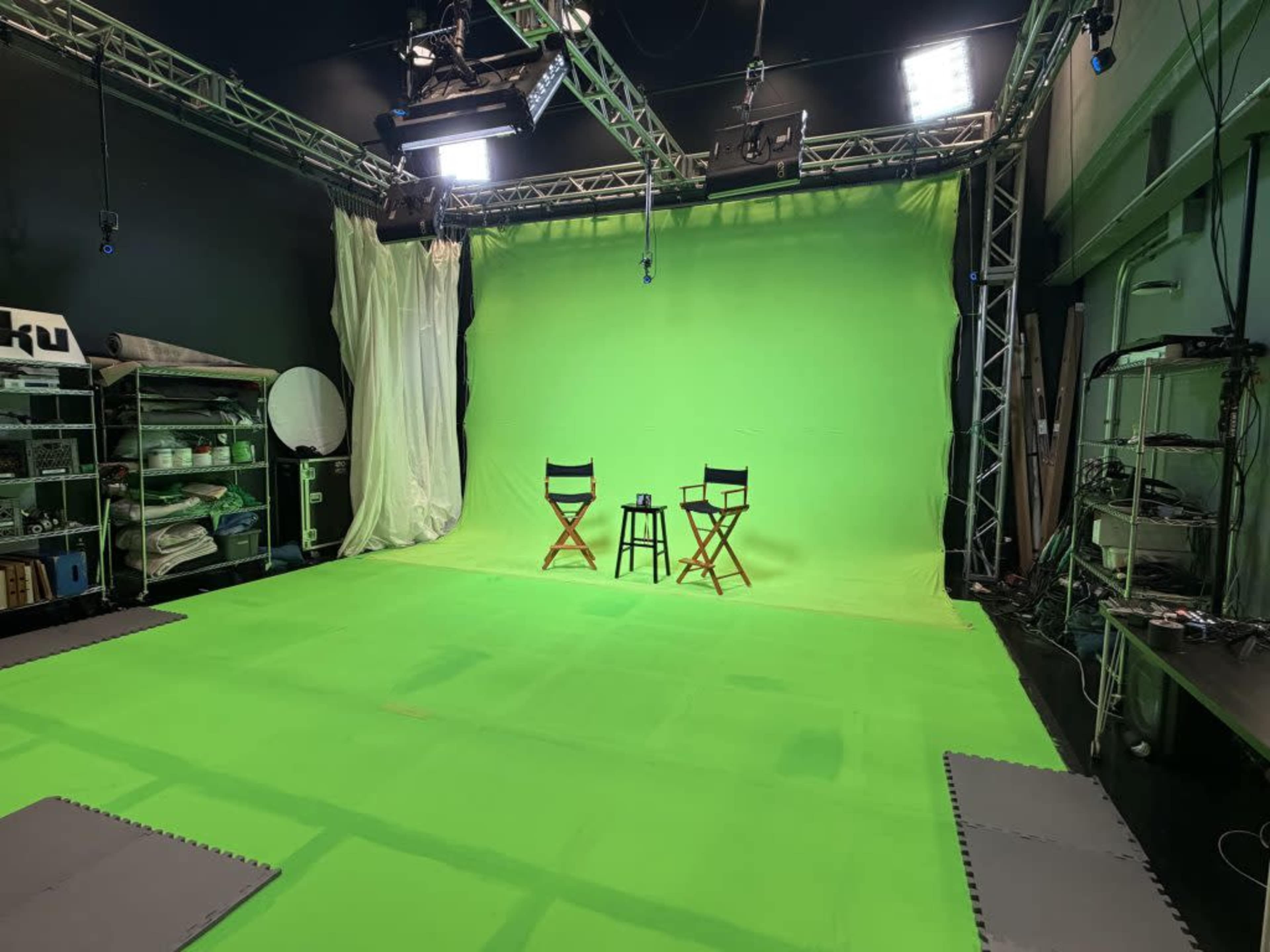 The image shows a green screen studio setup with two director's chairs positioned in front of a large green backdrop, surrounded by lighting equipment and various props.