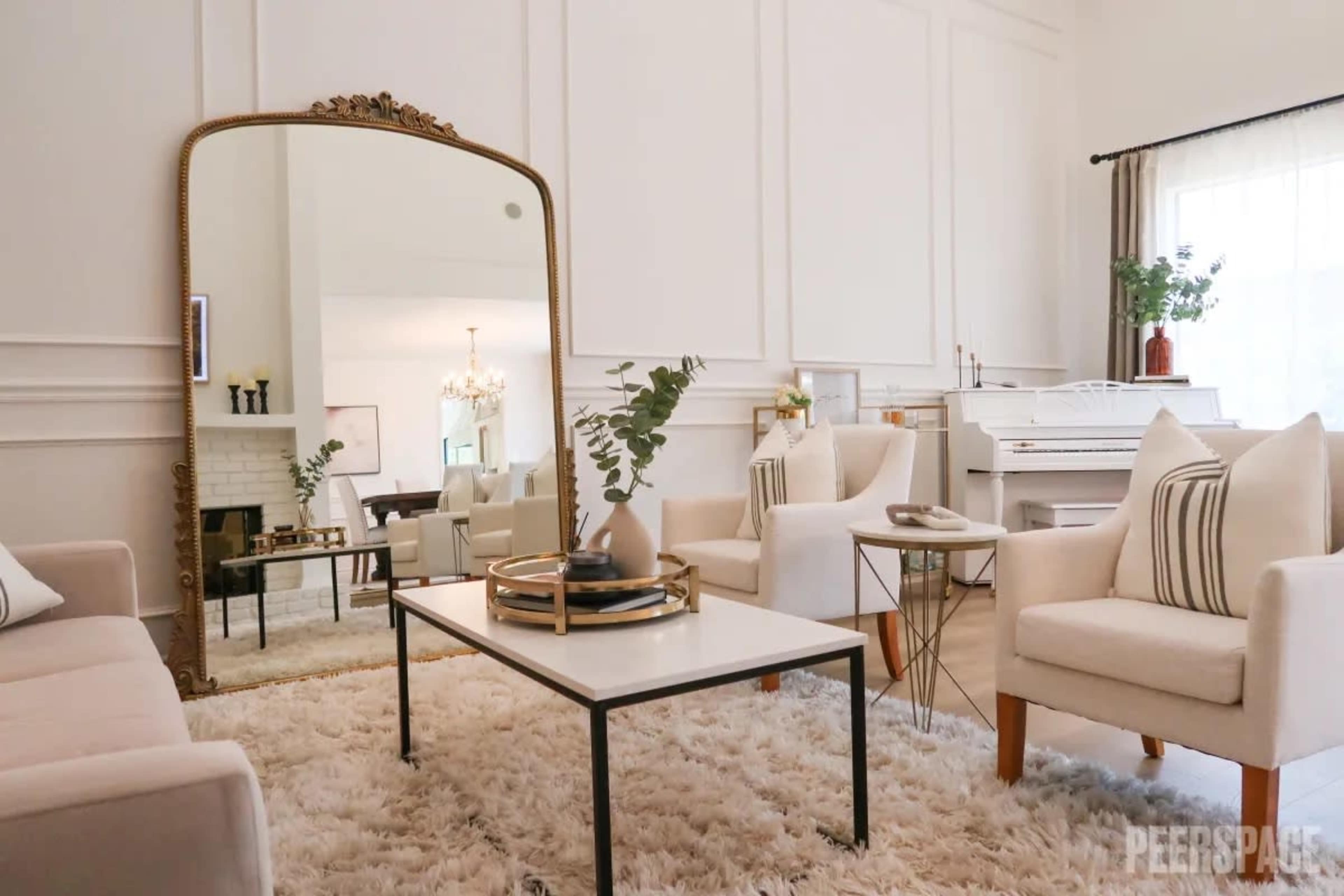 A modern living room with a large mirror, white sofas, a coffee table, and a decorative rug, featuring light-colored walls and natural light from nearby windows.