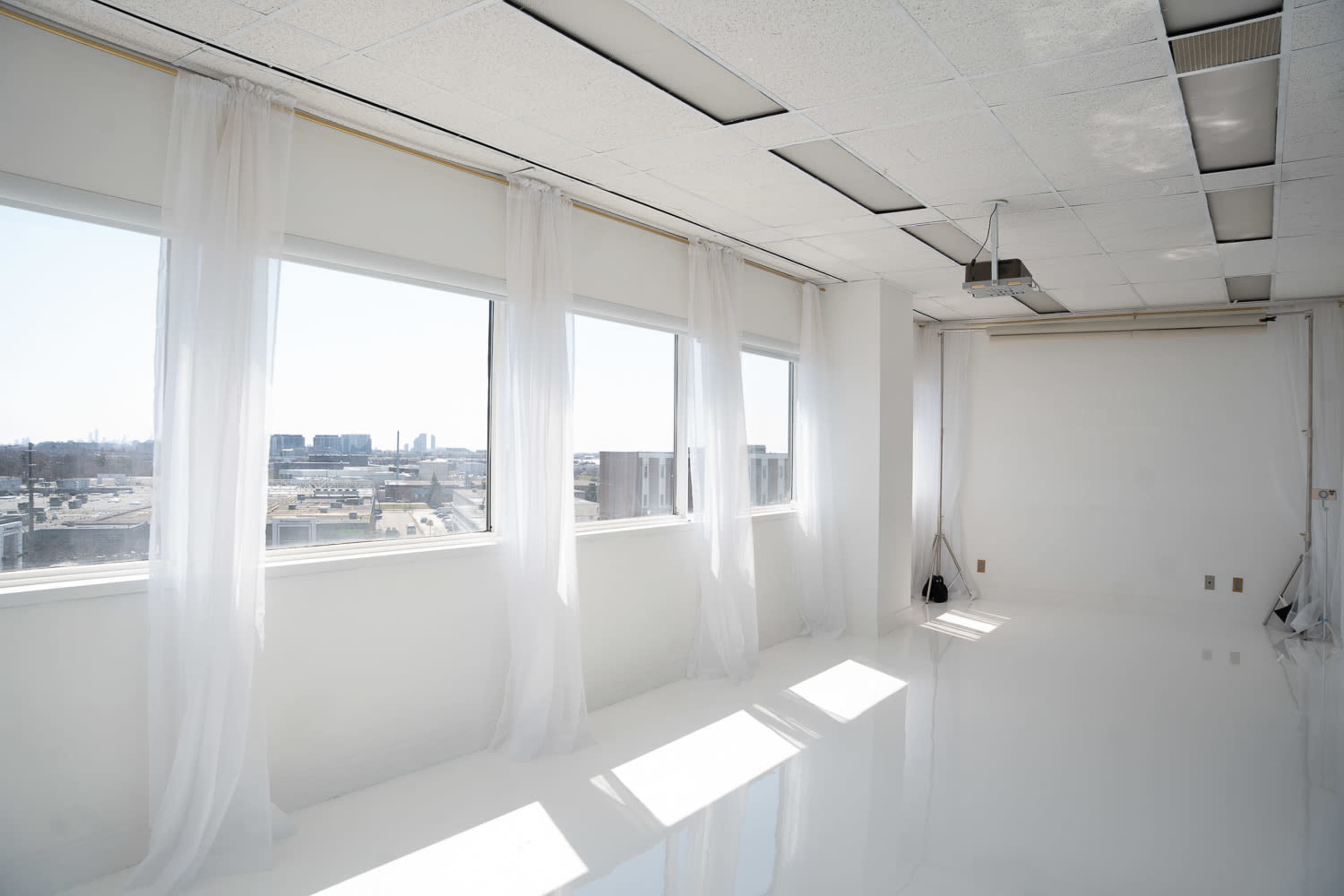 The image shows a bright, empty room with large windows dressed in sheer curtains, revealing a cityscape in the background.