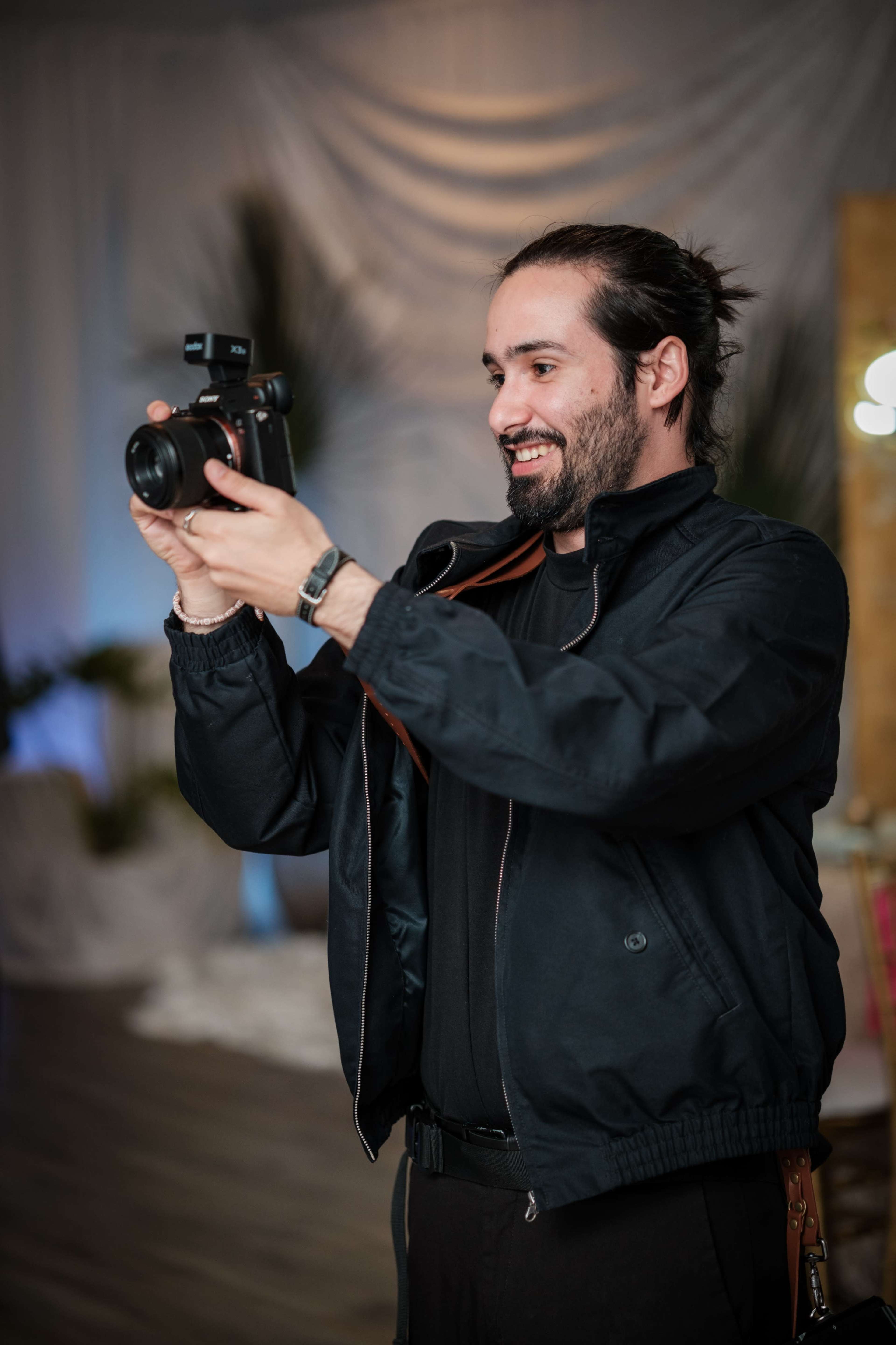 A man holds a camera and smiles while taking a photograph at an event.
