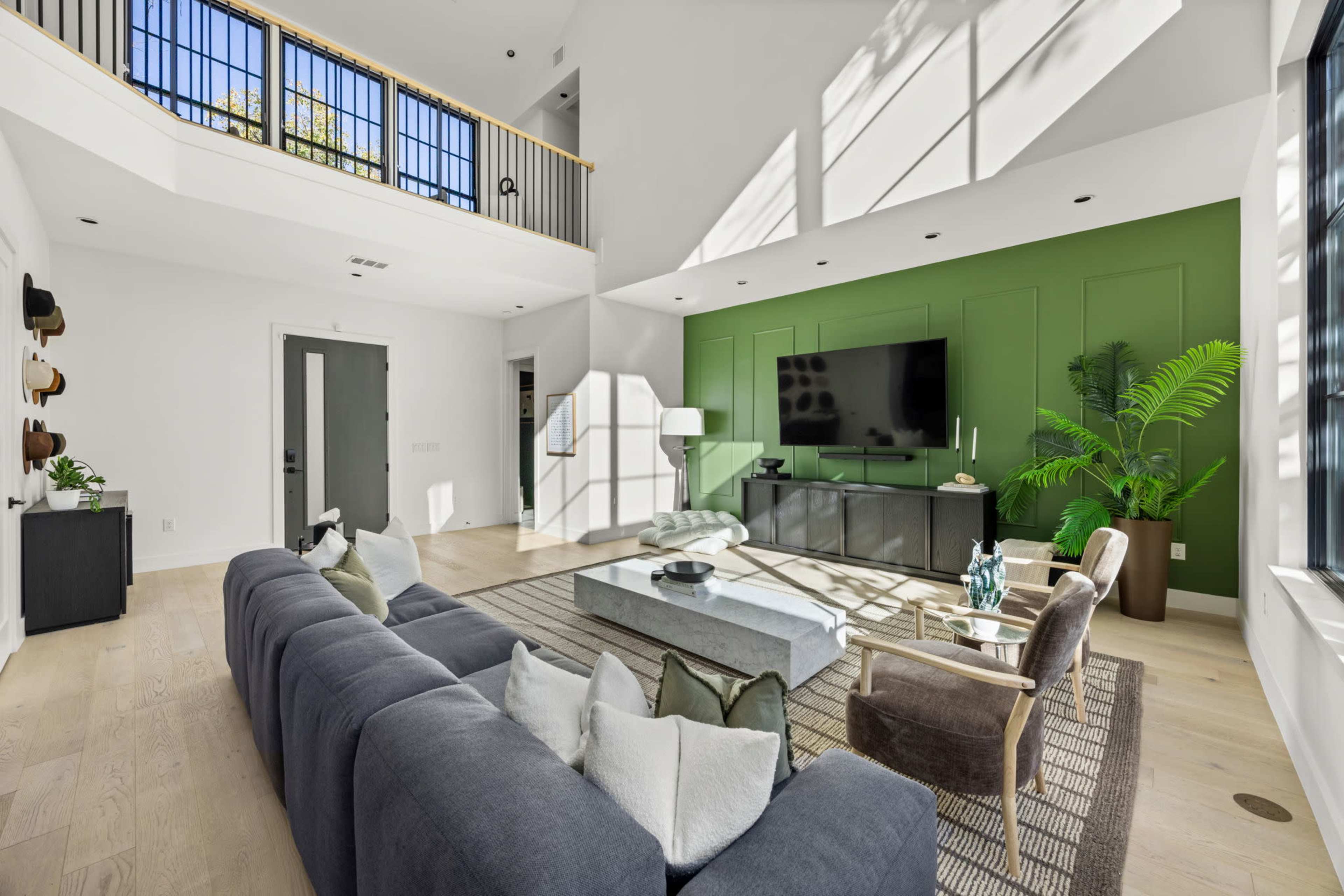 The image shows a modern living room with high ceilings, large windows, a sectional sofa, a coffee table, and a green accent wall featuring a television.