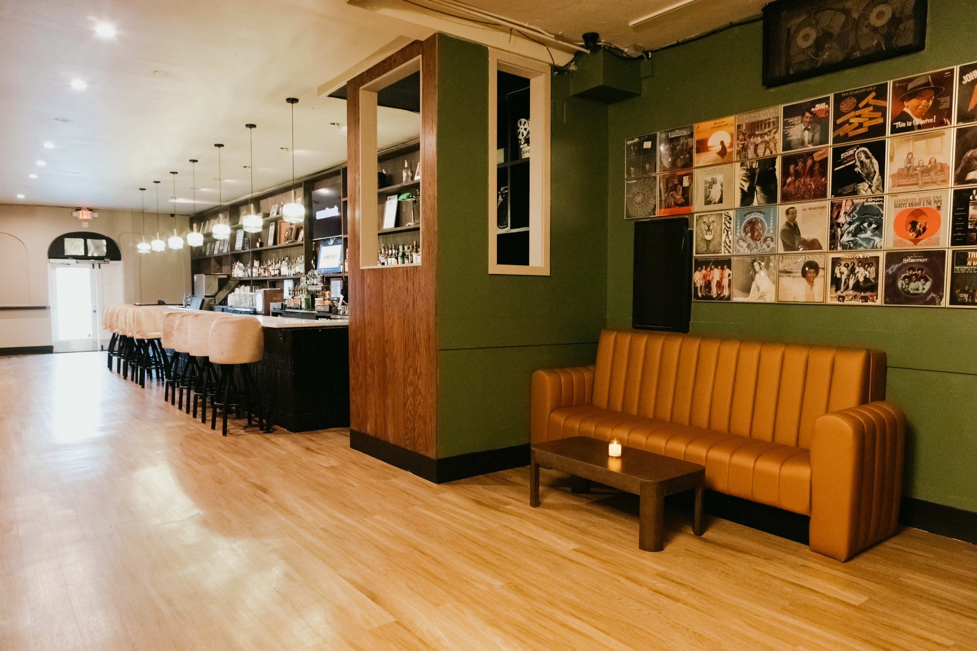 The image shows a cozy bar interior with a brown leather sofa, wooden coffee table, and a wall adorned with vinyl records, alongside a well-lit bar area featuring stools.