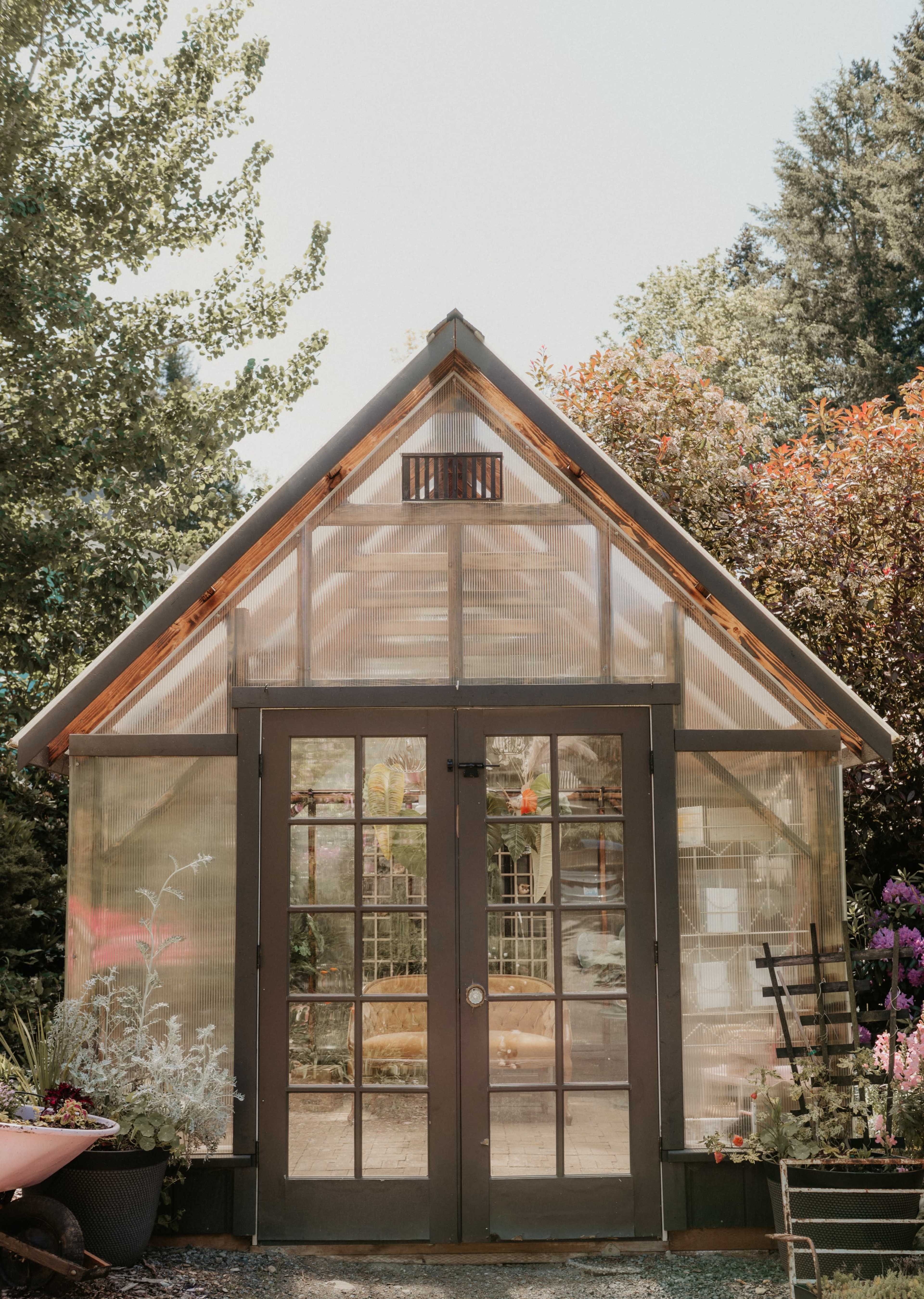 A glass-paneled greenhouse with a wooden frame and double doors is surrounded by various plants and flowers.