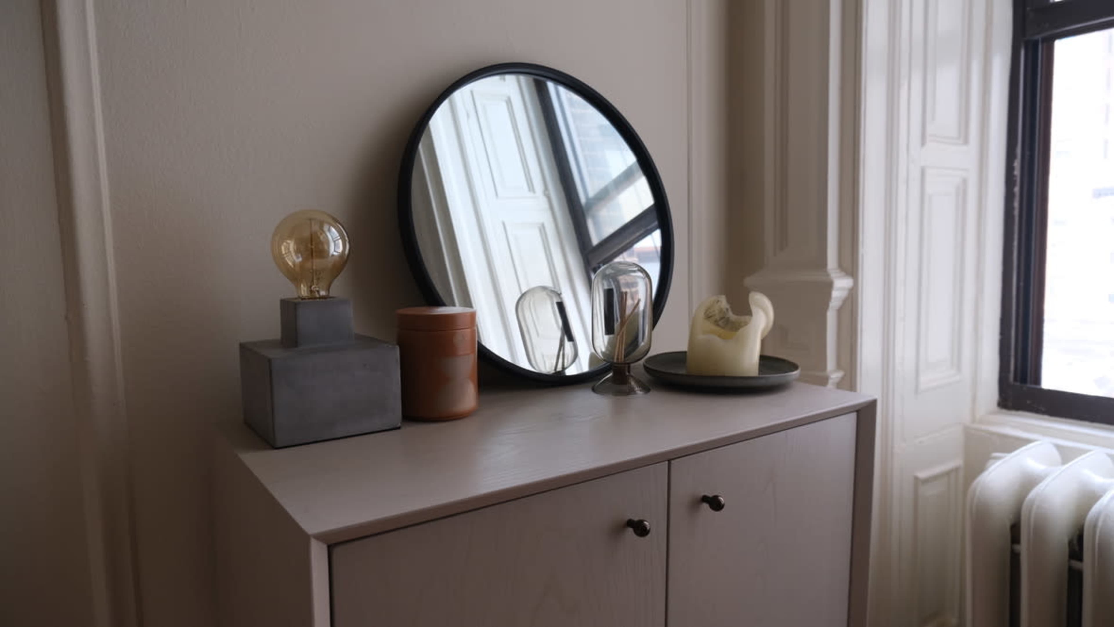 A round mirror and decorative items are arranged atop a light-colored cabinet near a window with a view.