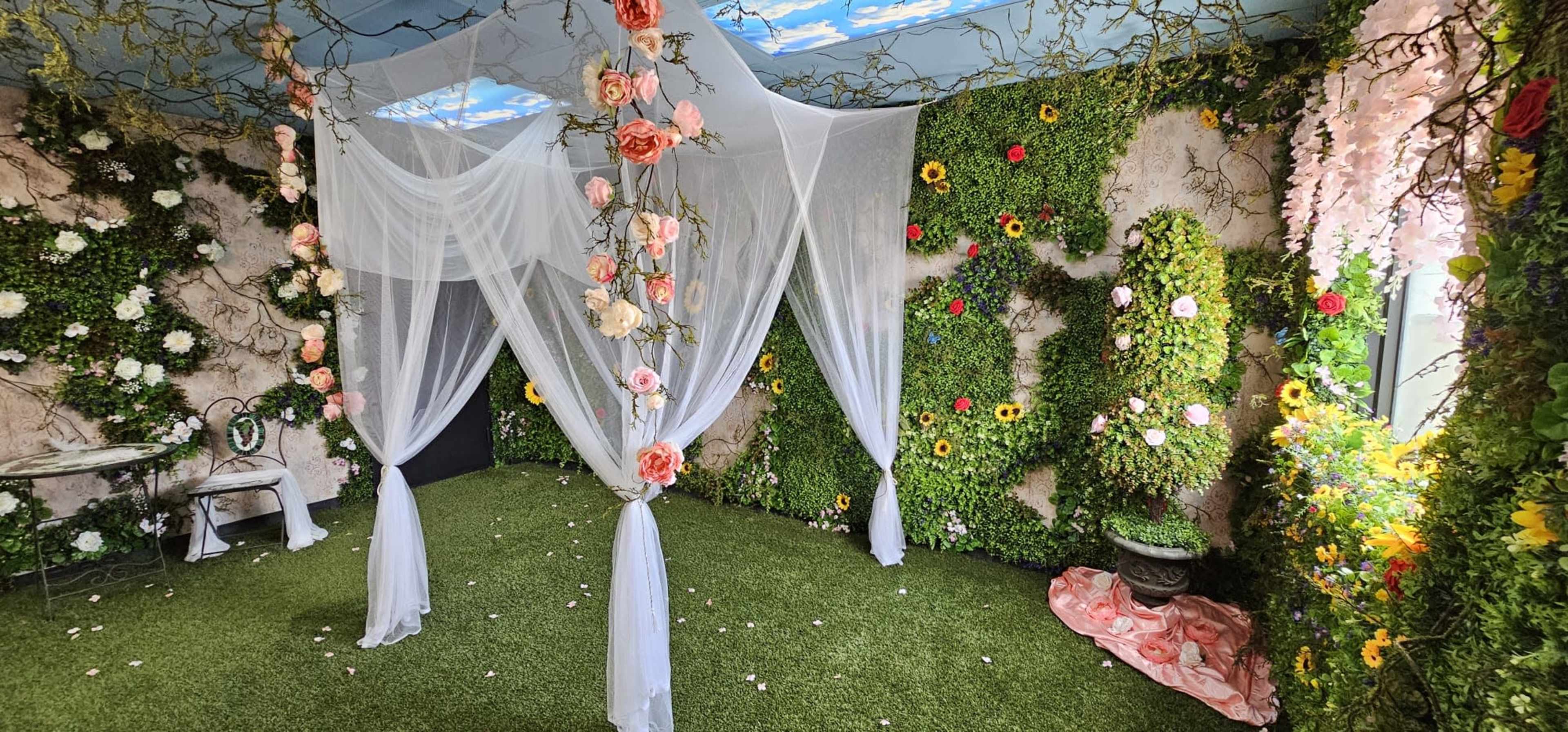 A decorated room featuring fabric drapery, artificial flowers, and greenery on the walls, with a grassy floor and a small table.