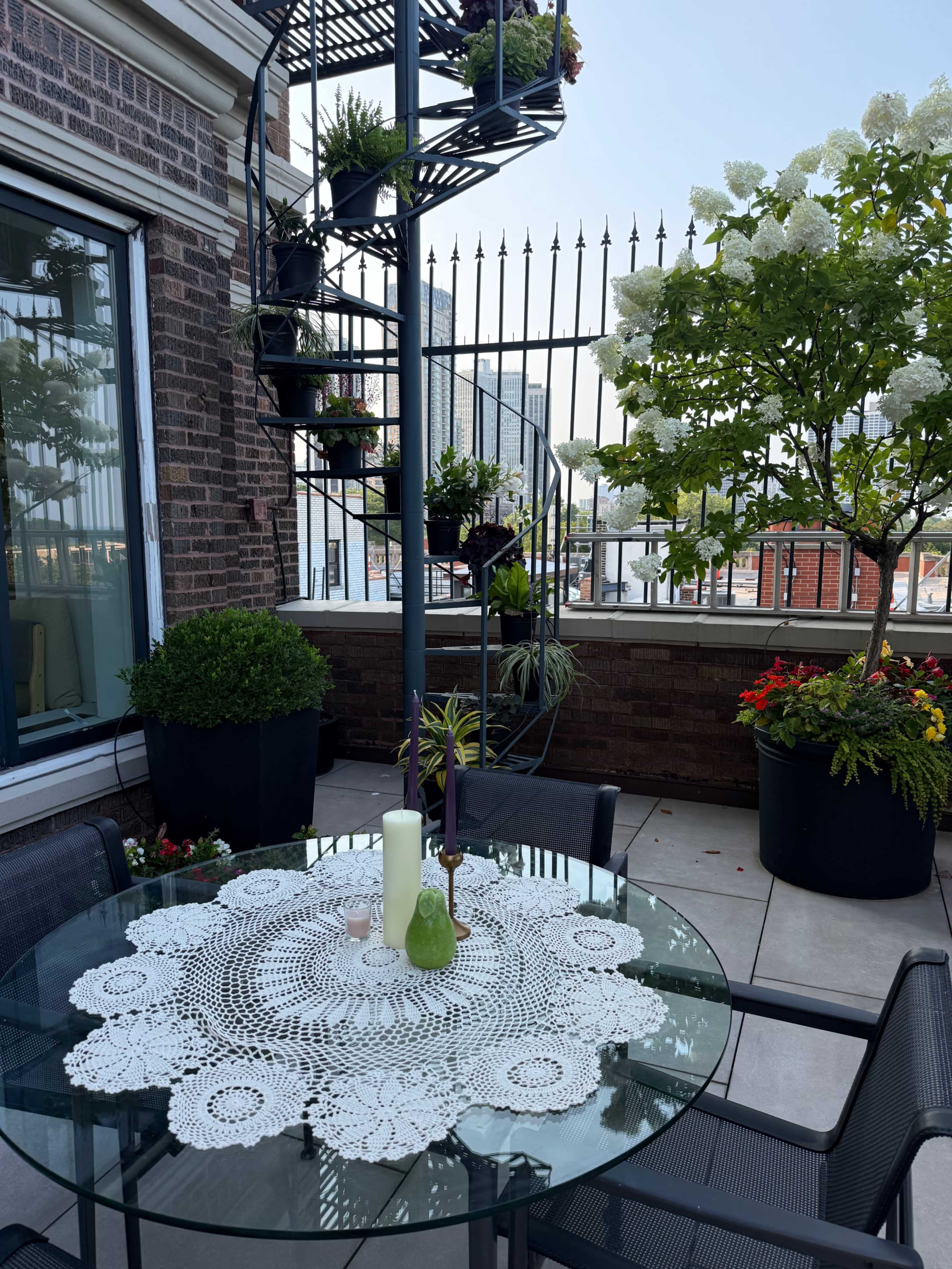 The image shows a patio with a glass table set with a lace tablecloth, surrounded by potted plants and a spiral staircase leading to a rooftop area.