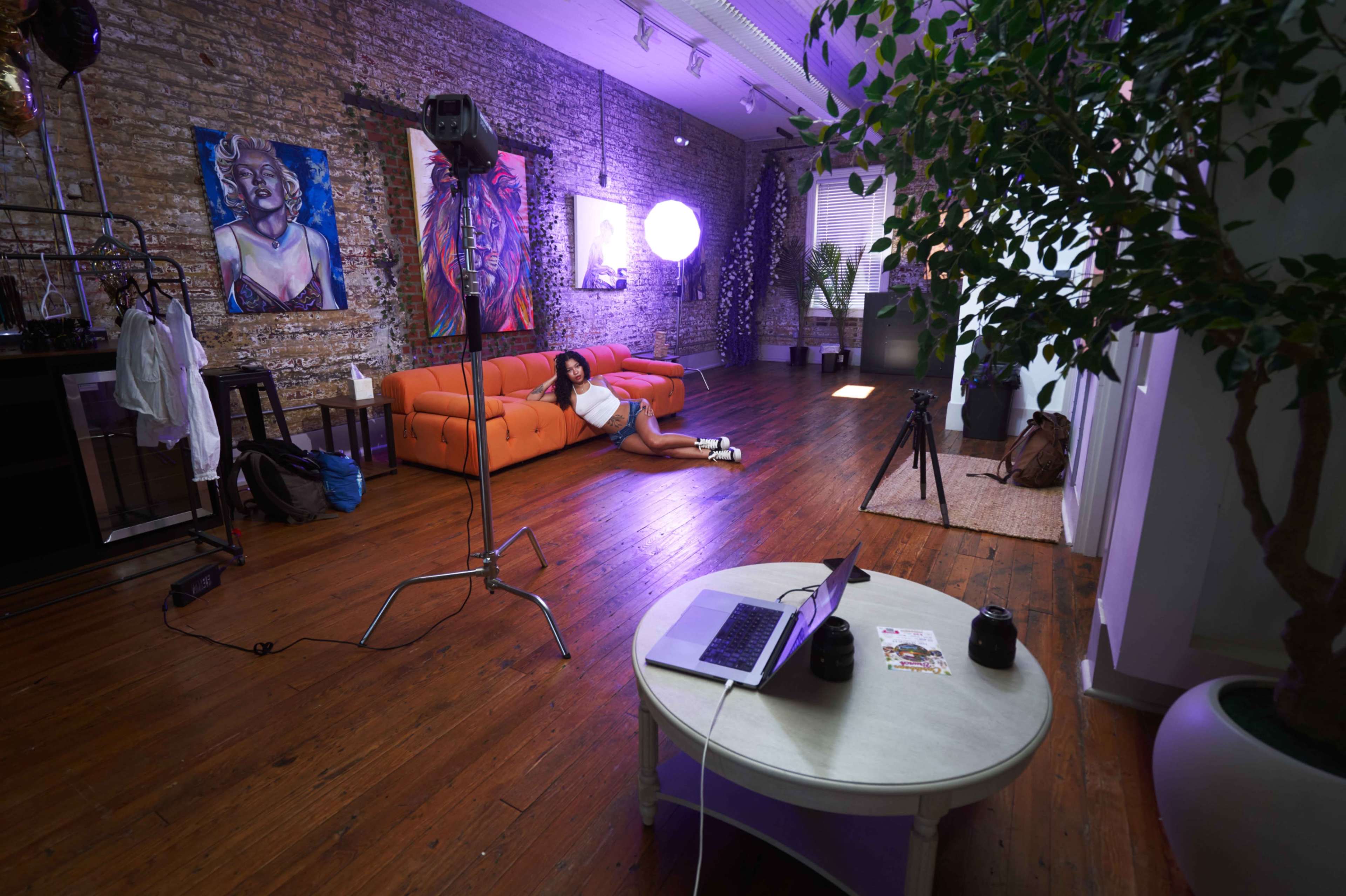 A photo studio features an orange sofa, several art pieces on the wall, and a model lounging on the floor near a laptop and various lighting equipment.