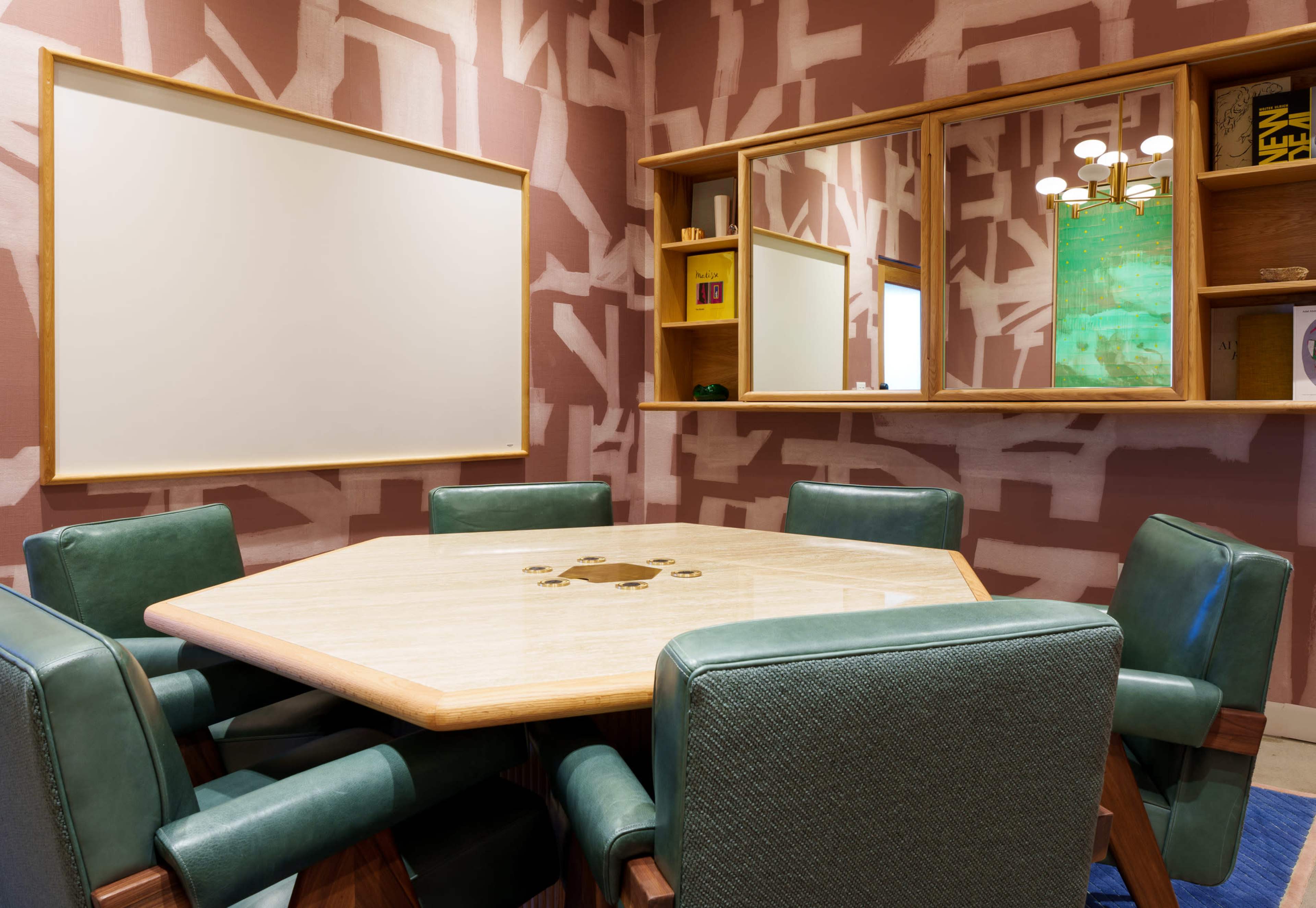 A well-furnished meeting room features a hexagonal table surrounded by green chairs, with a large blank whiteboard and a mirror on the wall.