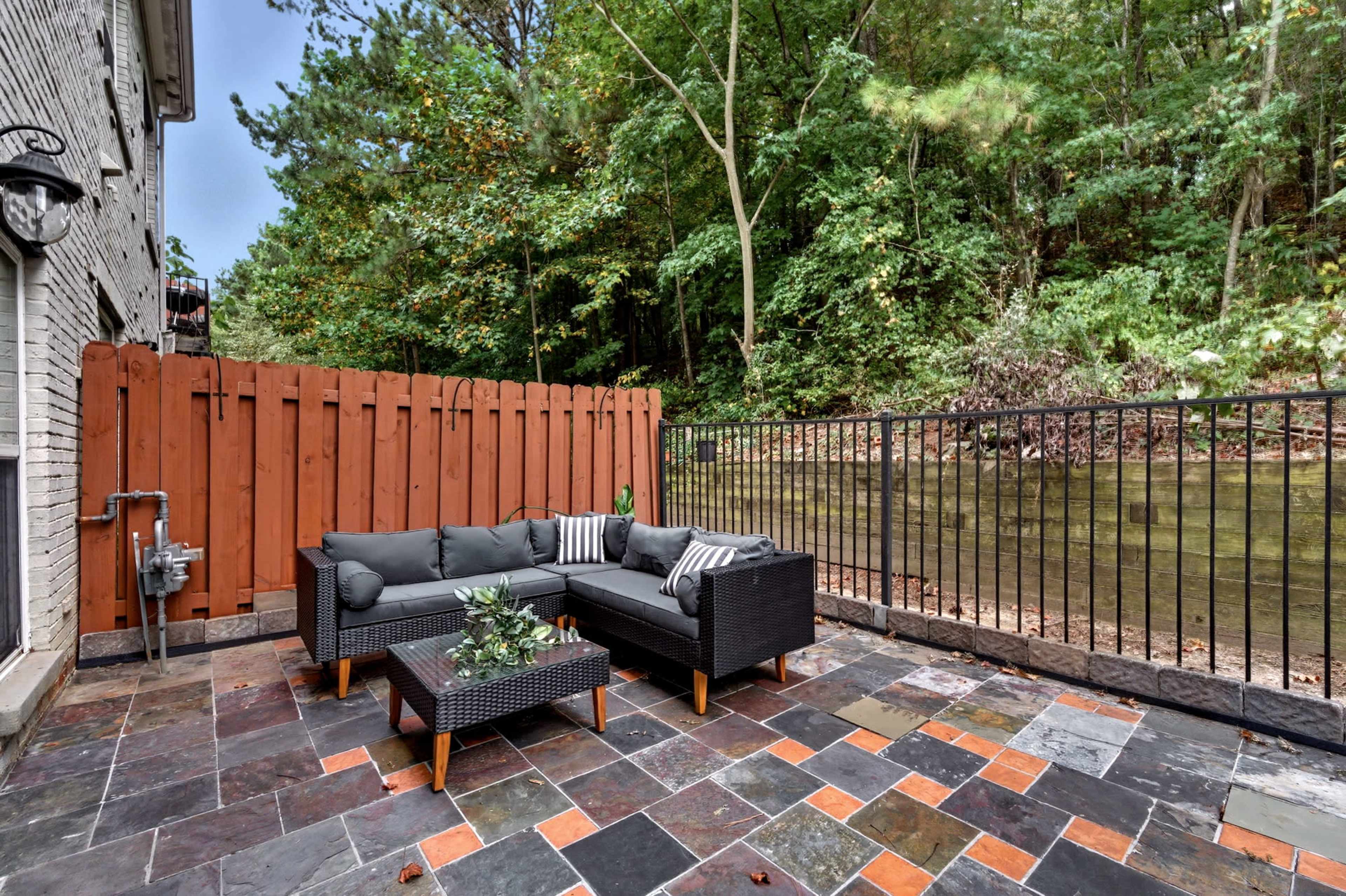 The image shows a patio area with a dark sectional sofa and a coffee table, surrounded by a wooden fence and a backdrop of trees.