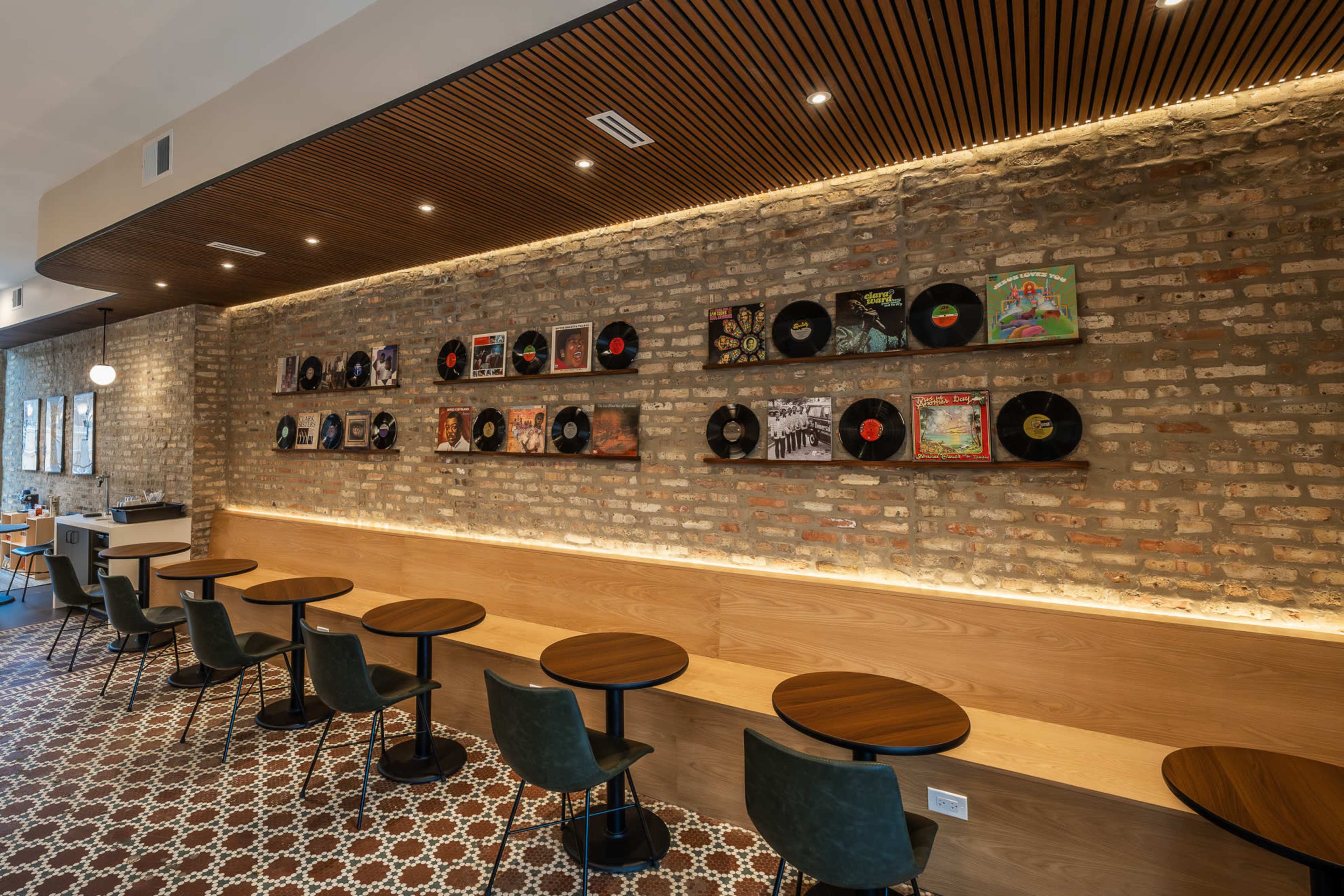 The image shows a modern café interior featuring a brick wall adorned with vinyl records, wooden seating along the wall, and small round tables.