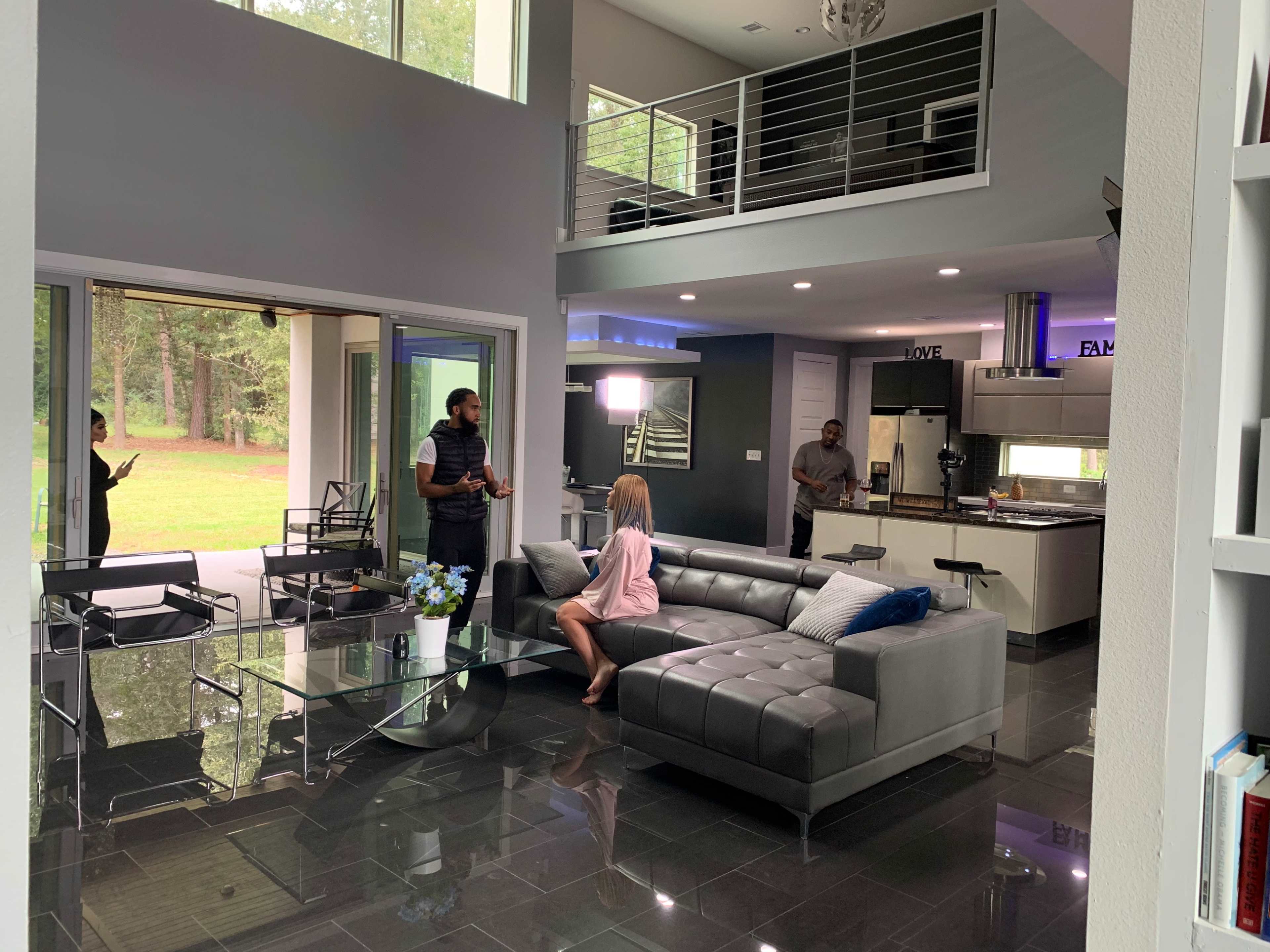 A modern living room features a woman sitting on a gray couch while two men stand nearby, with large windows revealing a view of a forested area outside.