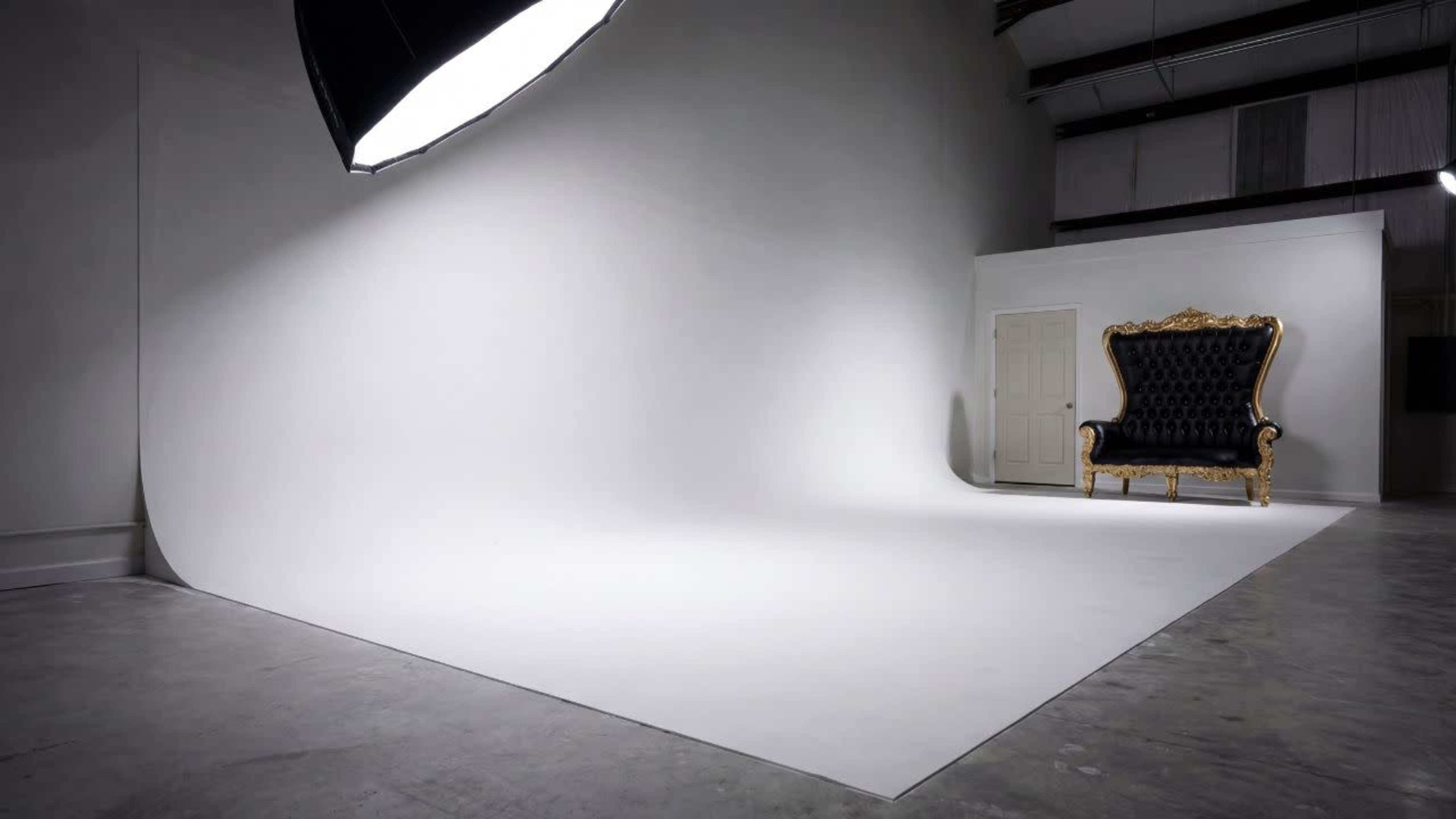 The image shows a spacious photography studio with a seamless white backdrop, a large lighting fixture, and an ornate black and gold throne positioned against a wall with a closed door.