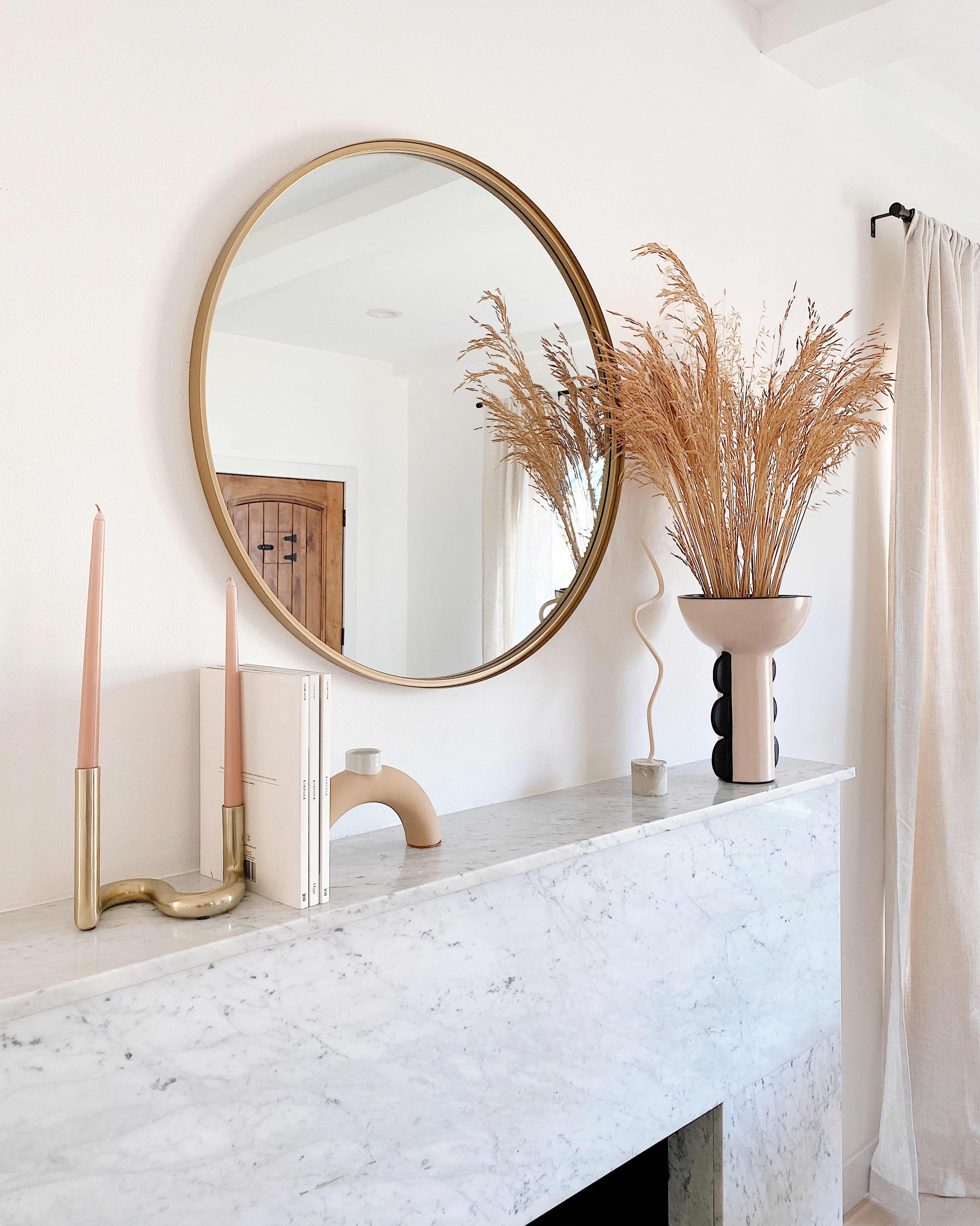 A round mirror hangs above a marble mantel adorned with candles, books, and a vase of dried grasses.