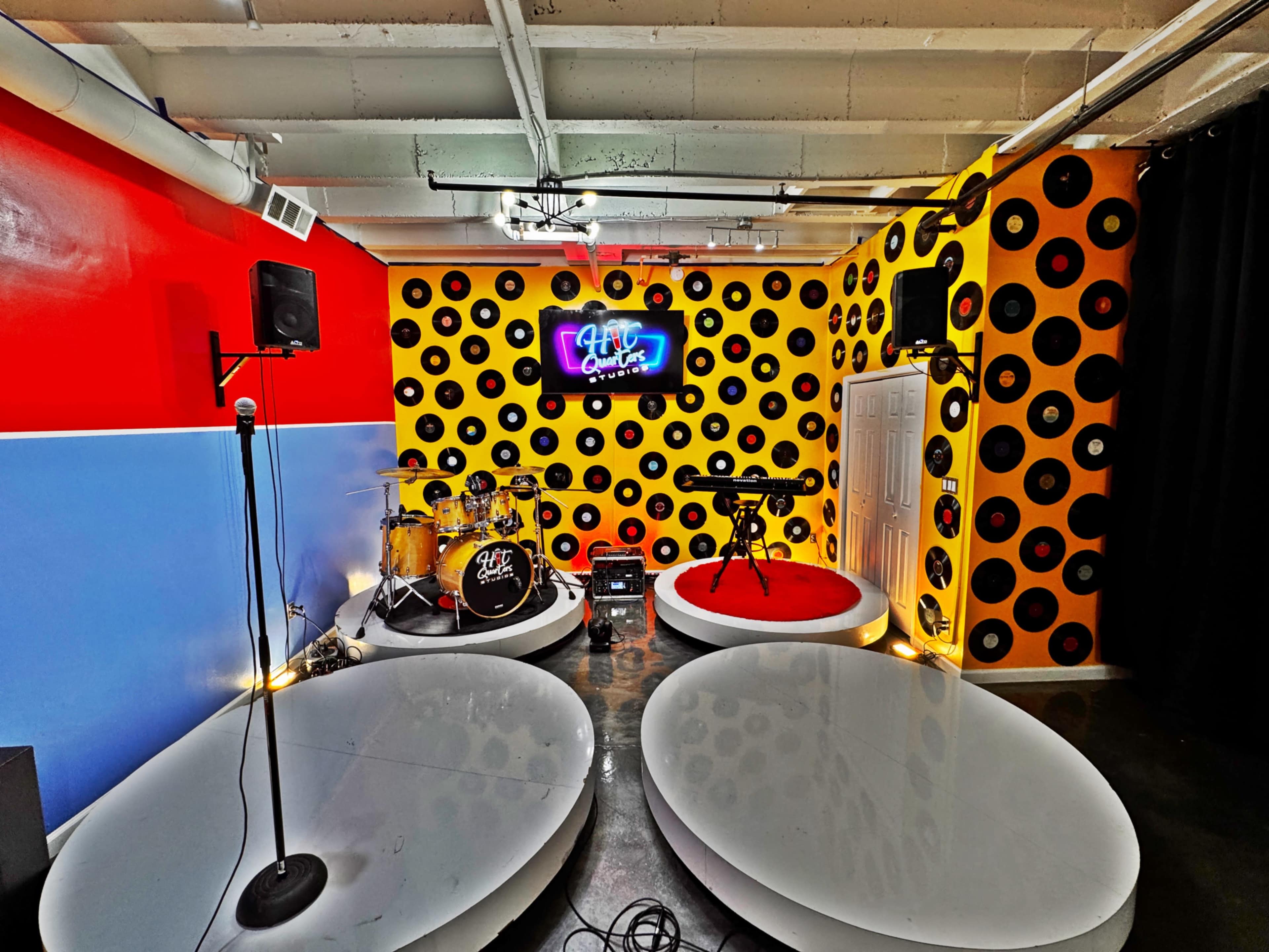 The image shows a vibrant music rehearsal space featuring a drum set, keyboard, microphone stand, and walls decorated with vinyl records.