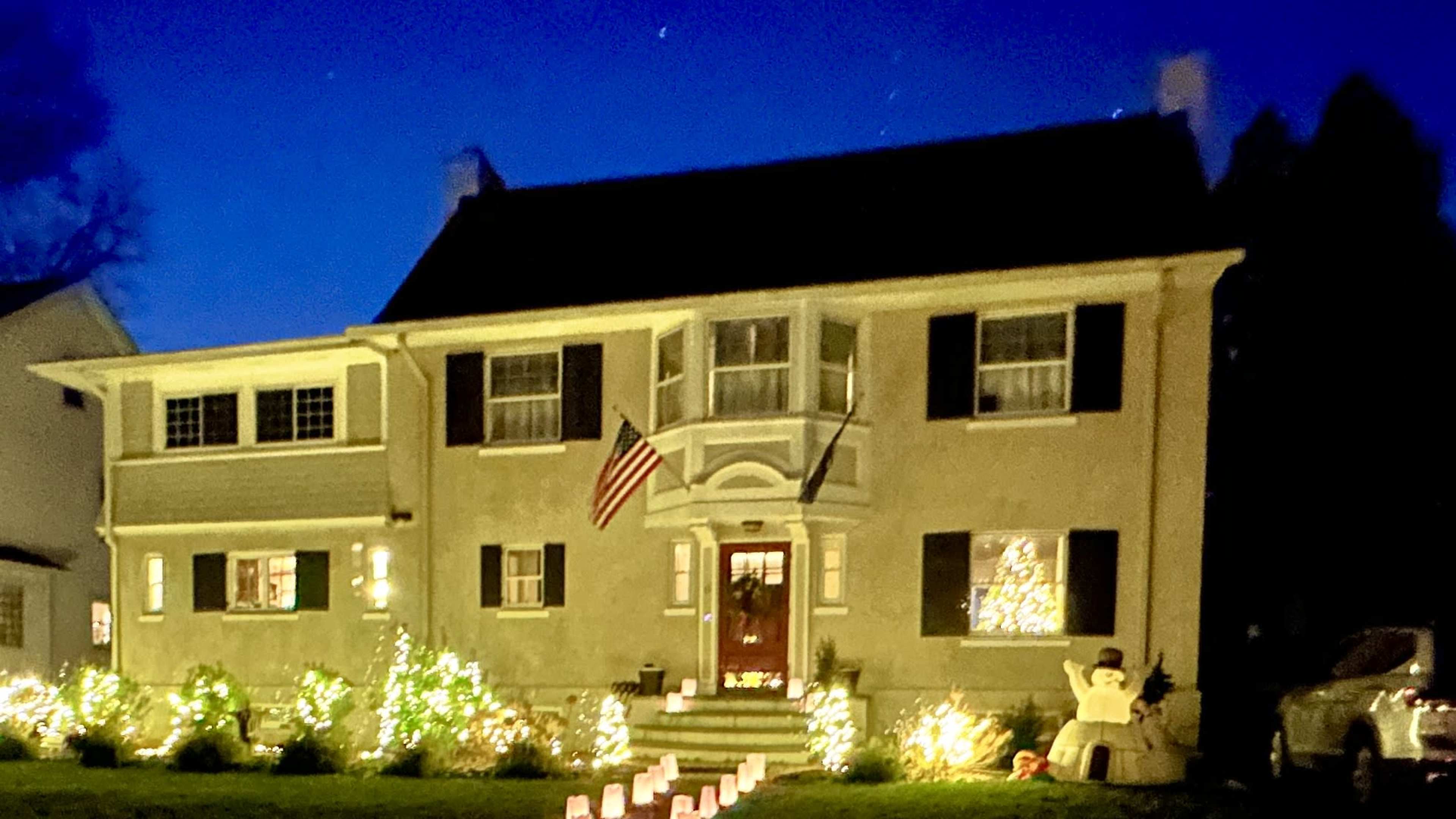 A two-story house with Christmas lights, an American flag, and a snowman decoration is illuminated against a twilight sky.