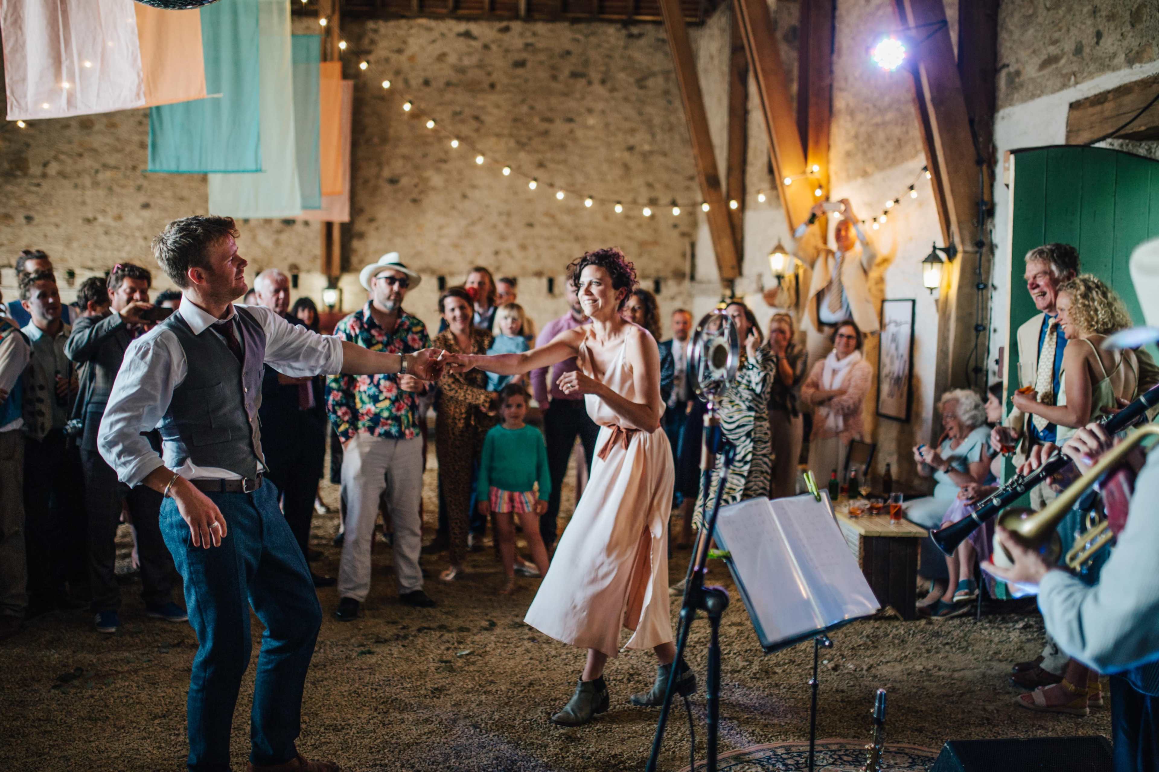 A couple dances in a rustic venue filled with guests, while a band plays music in the background.
