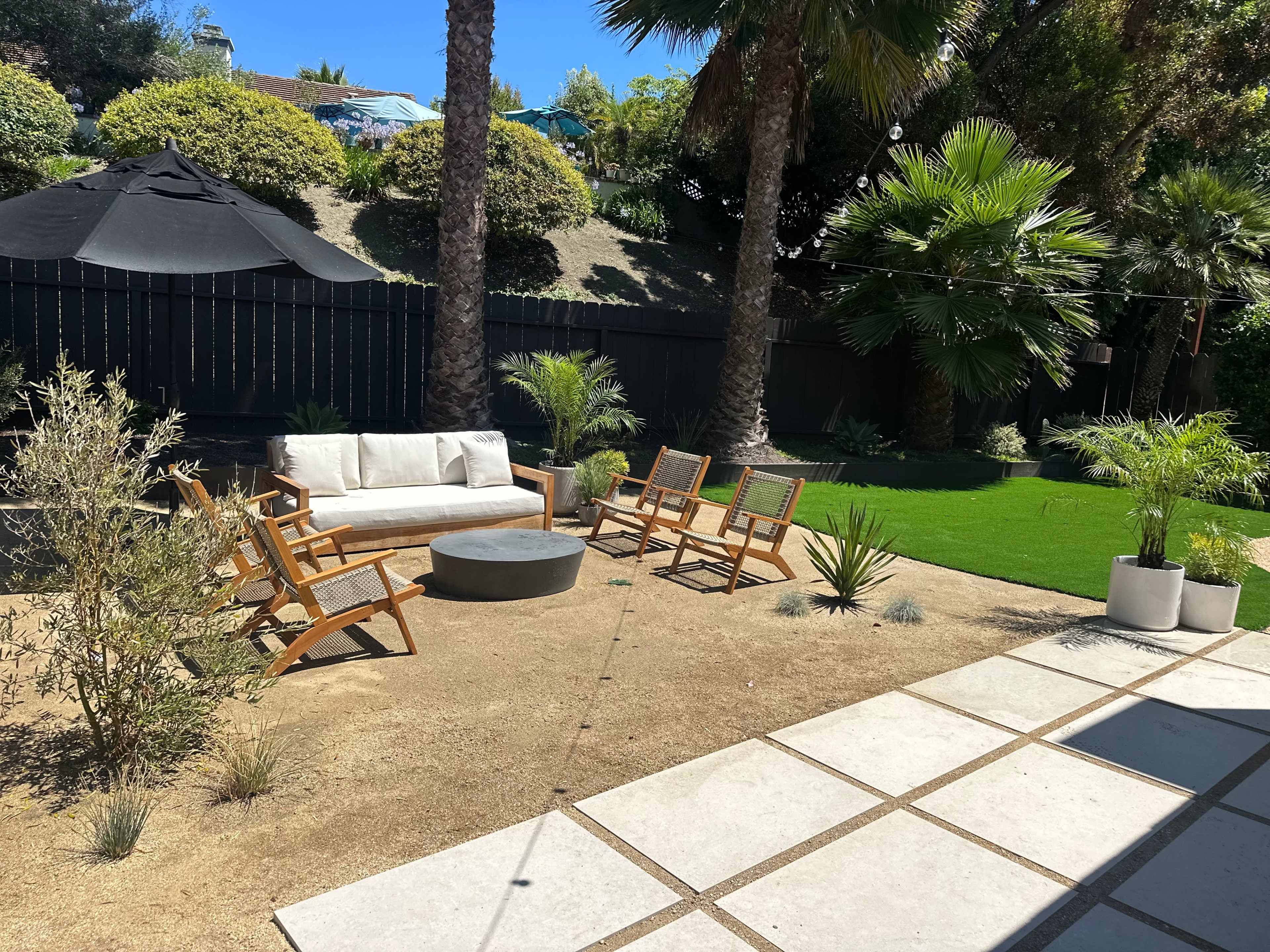 A landscaped outdoor area with a seating arrangement consisting of a sofa and wooden chairs, surrounded by plants and a circular coffee table on sandy ground.