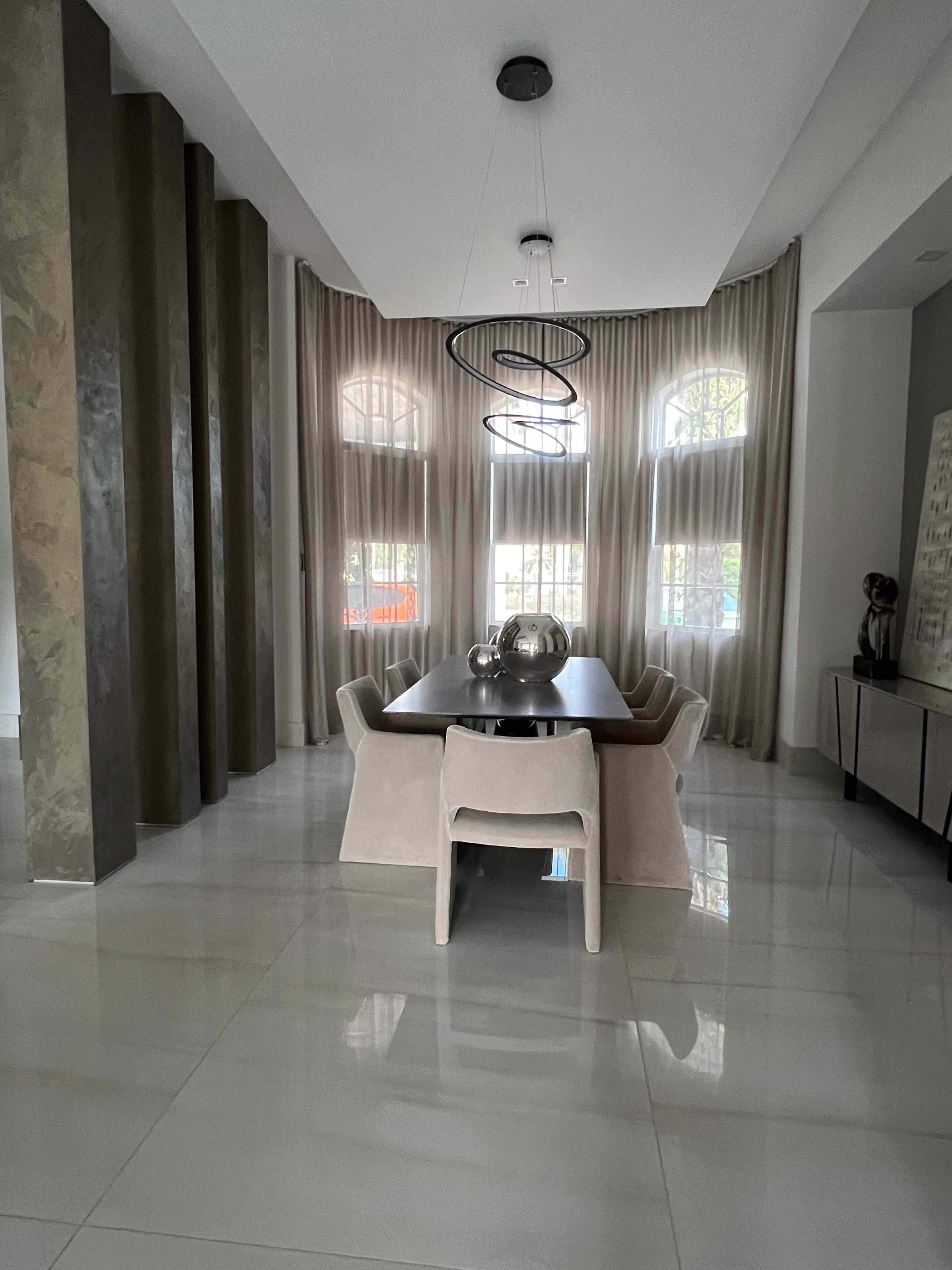 A modern dining room features a large table surrounded by light-colored chairs, tall windows with sheer curtains, and a geometric pendant light overhead.