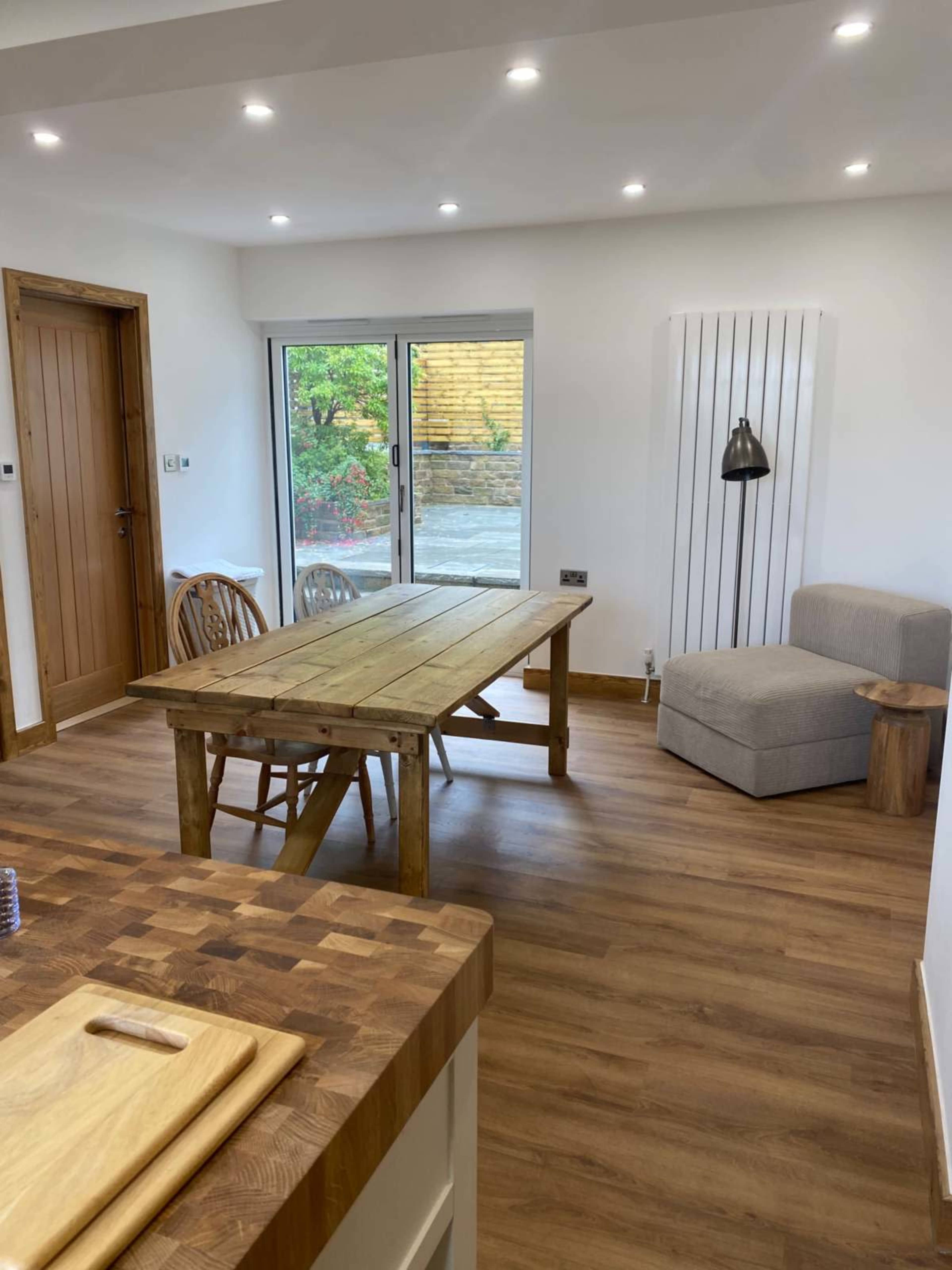 The image shows a modern kitchen-dining area with a wooden table, chairs, and a comfortable chair beside a large window overlooking a garden.