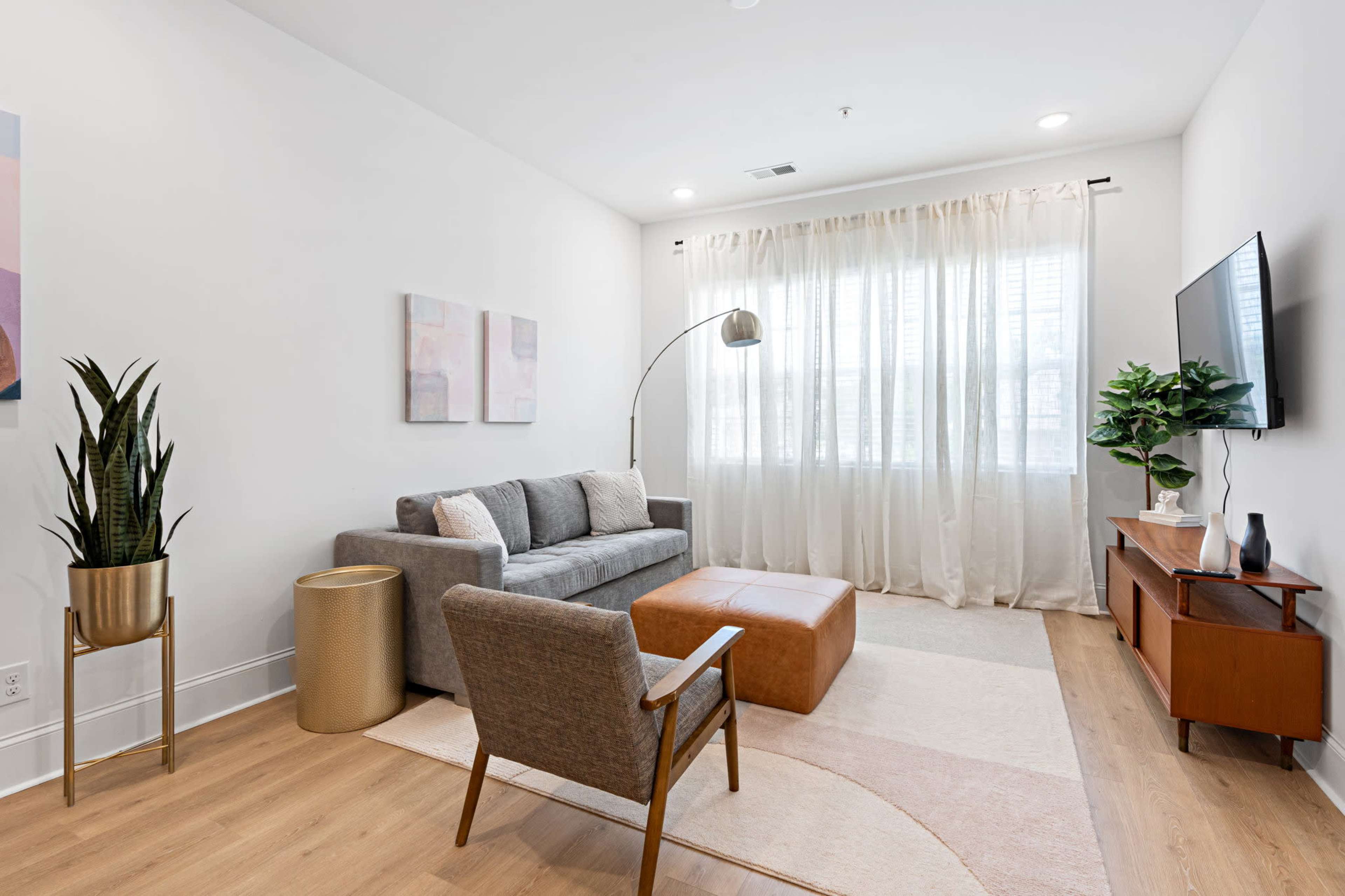 A modern living room with a gray sofa, a brown ottoman, a decorative chair, and large windows covered with light curtains.