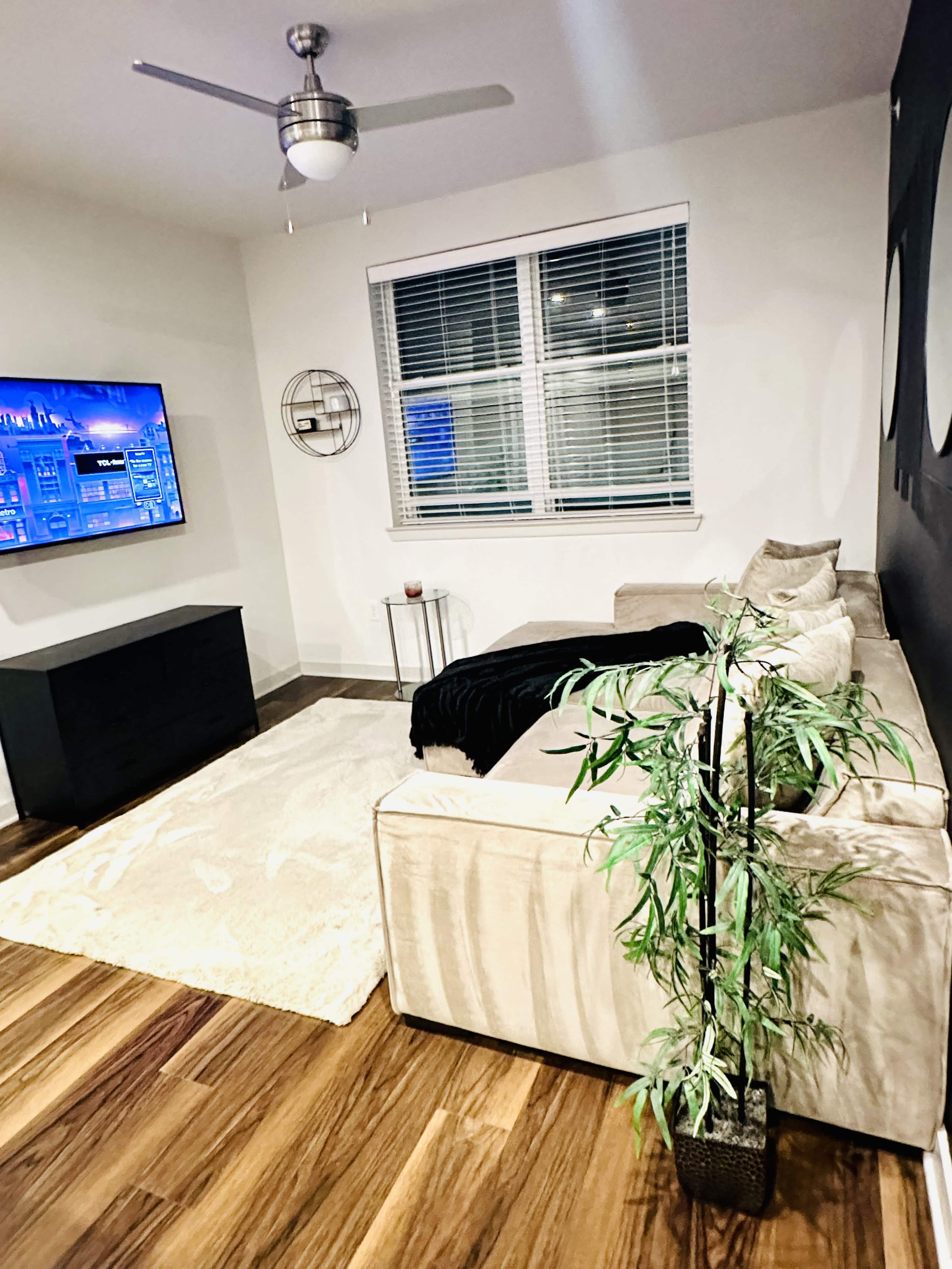 The image shows a modern living room with a beige sectional sofa, a television mounted on the wall, and a potted plant in the corner.